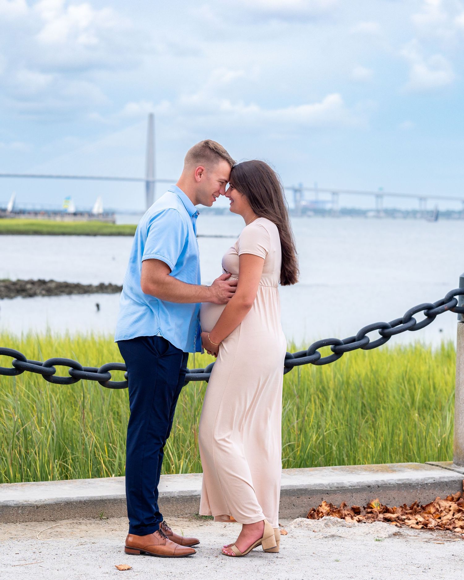 Husband and pregnant wife wearing white dress stand on pier facing each other