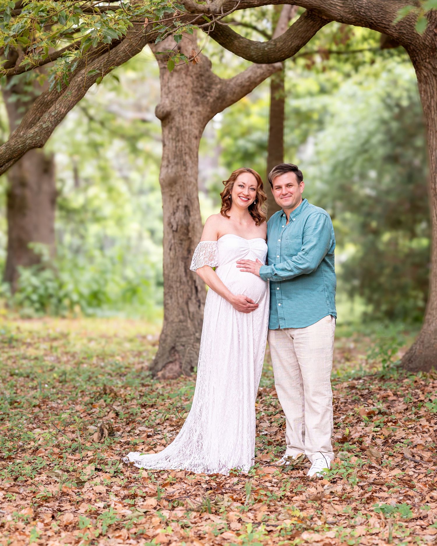 A tender maternity photoshoot in a wooded outdoor setting during autumn with fallen leaves on the ground.