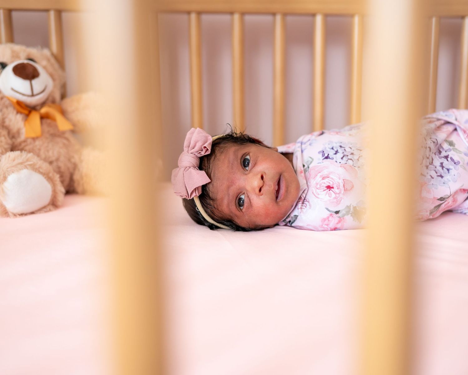Baby lying in white crib with teddy bear visible through wooden slats in pink outfit.