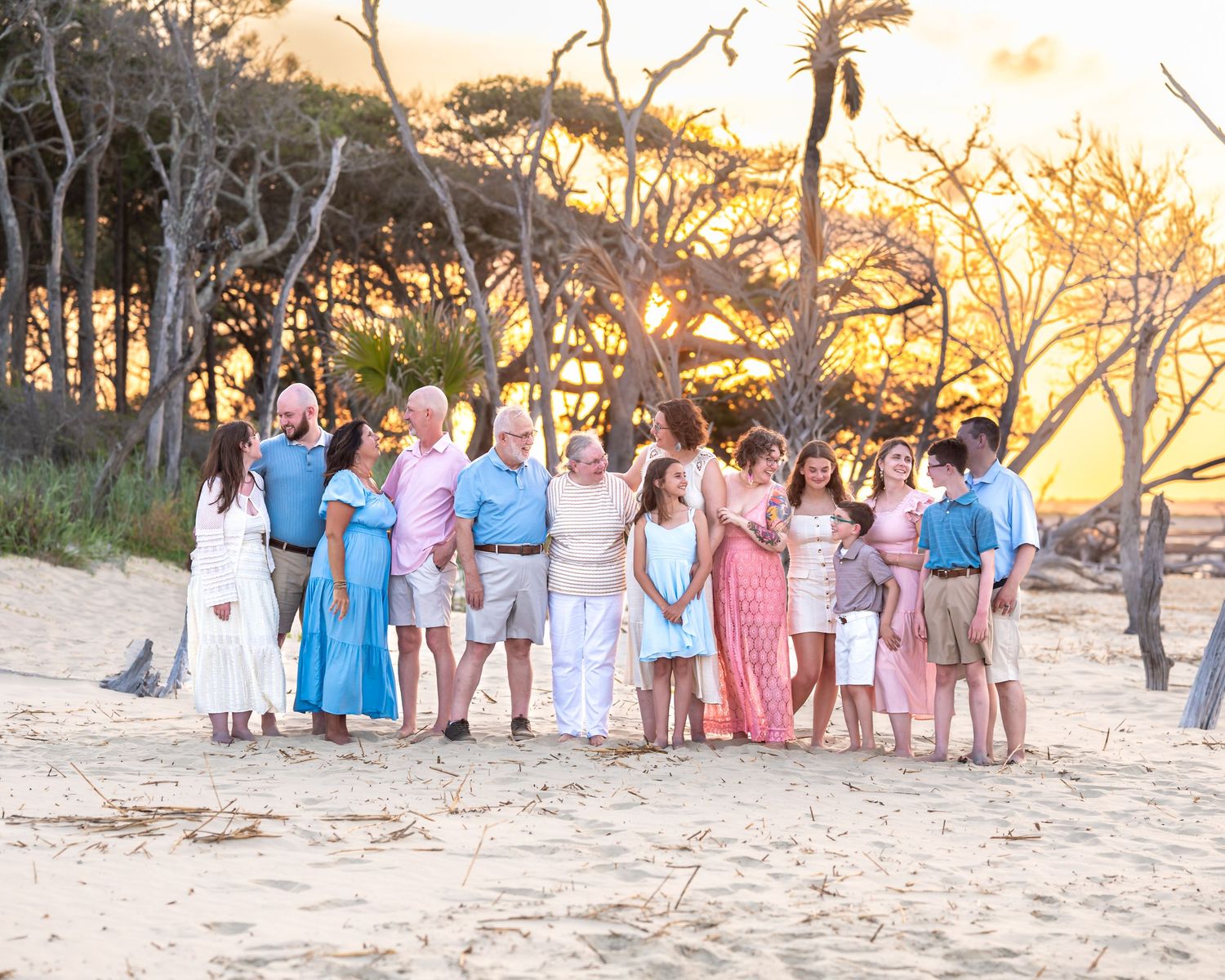 Large, extended family group posing together on sandy beach at golden sunset with bare trees in background.