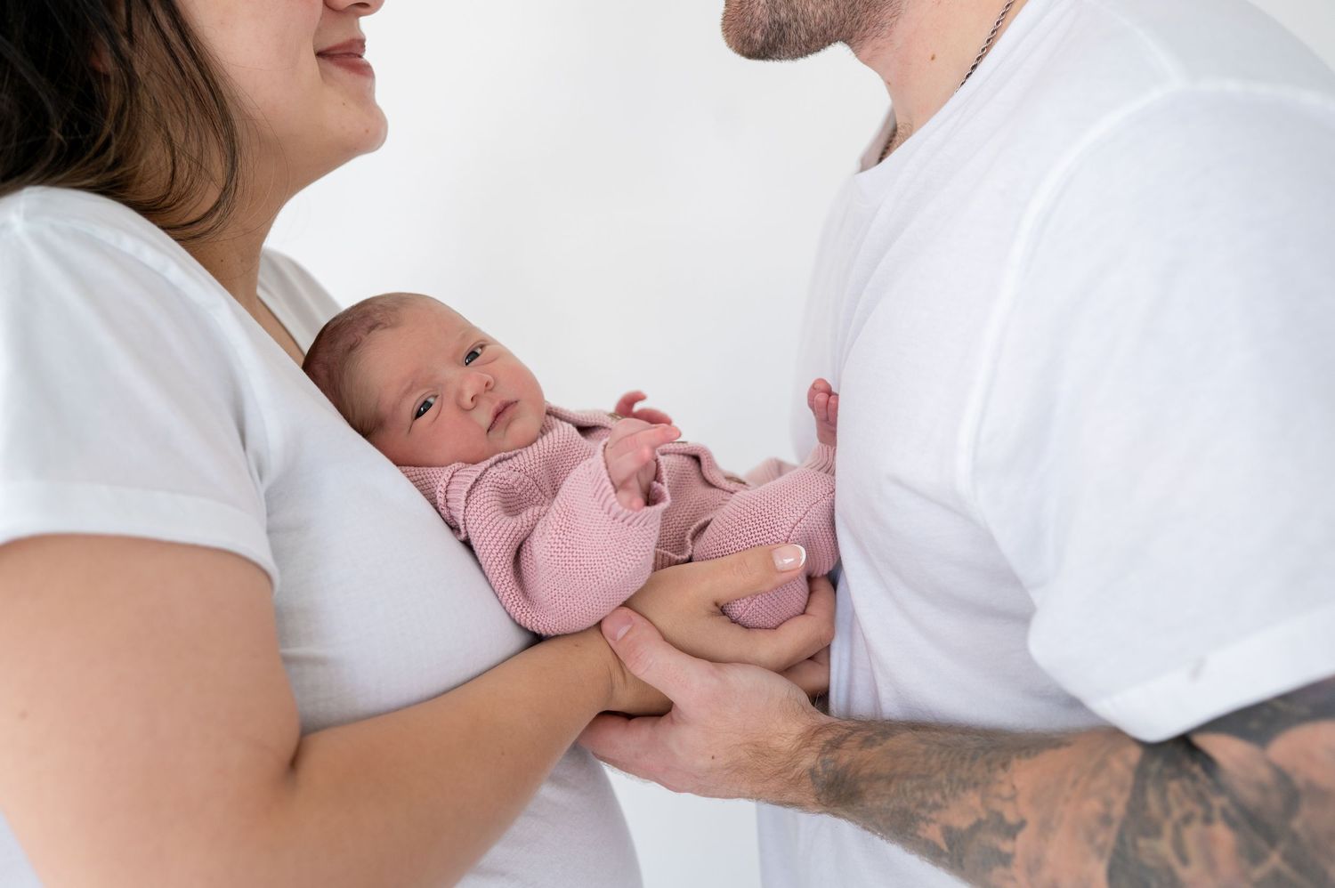 New parents in white shirts lovingly hold their newborn baby wrapped in a pink blanket against a white background.