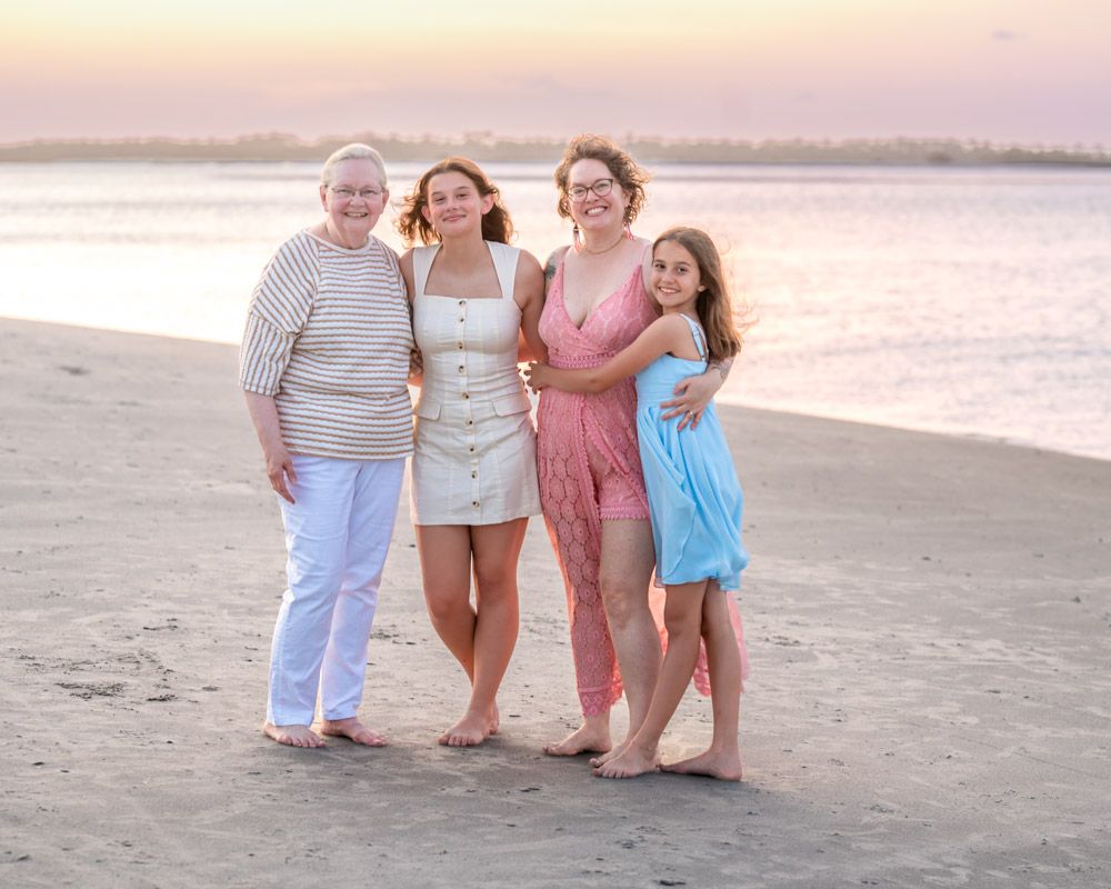 Four women pose together smiling on a sandy beach at sunset with calm water in the background.