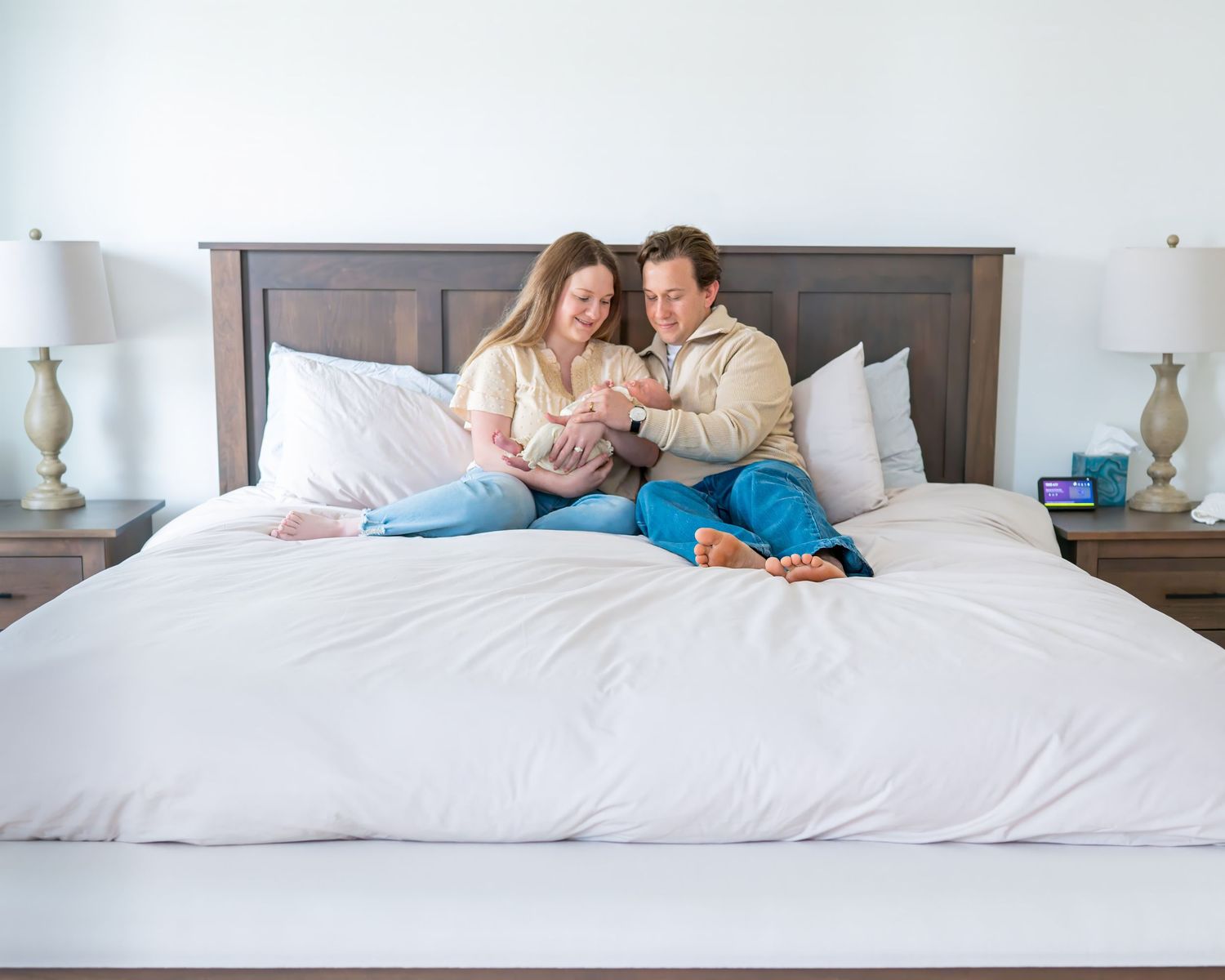 A couple sits together on a white bed with matching blue table lamps while looking at their newborn daughter