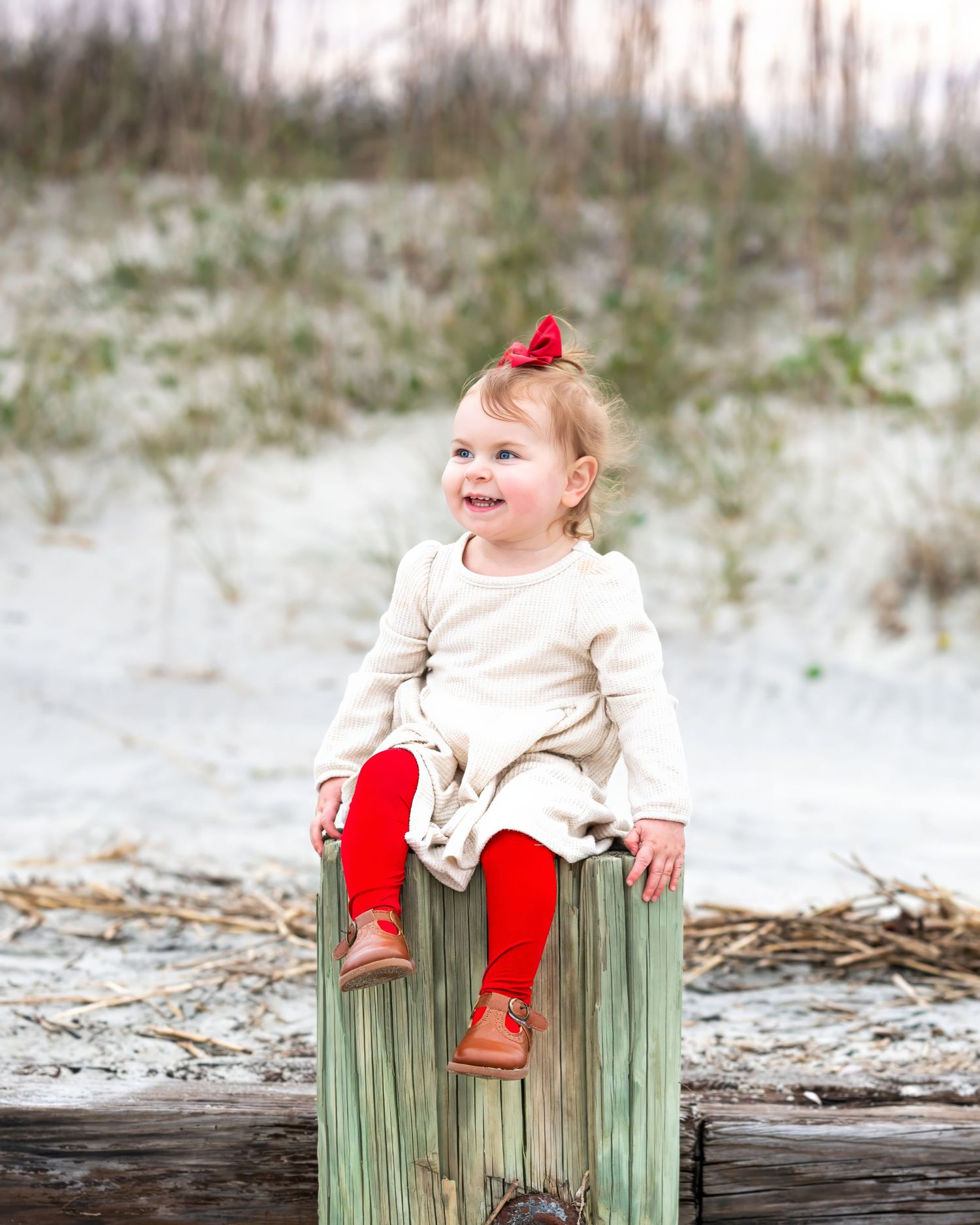 Child in cream dress and red tights sits happily on a weathered wooden post at the beach with sand dunes in background.