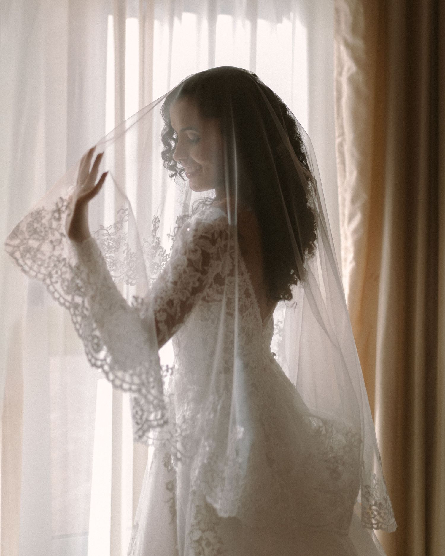 Bride in soft window light, framed through a doorway for an intimate, candid getting-ready moment.