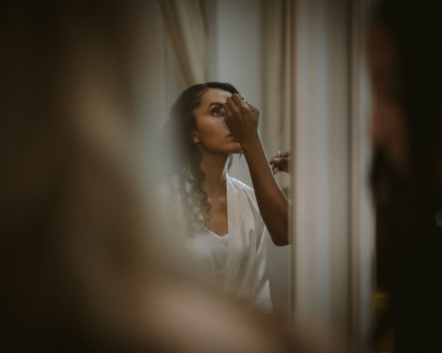 Bride applying makeup in soft window light, framed through a doorway for an intimate, candid getting-ready moment.