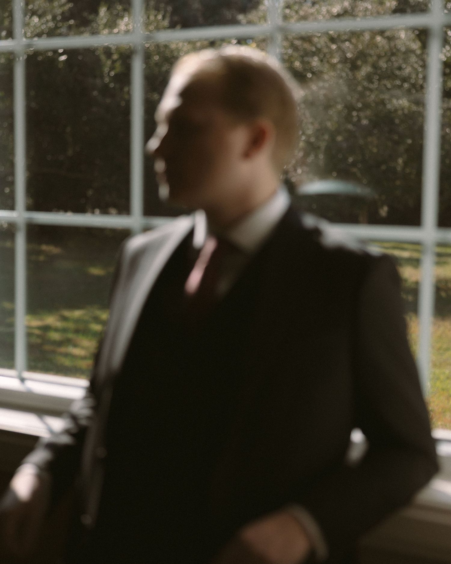 Groom standing by a window in natural light, a quiet, reflective moment before the ceremony.