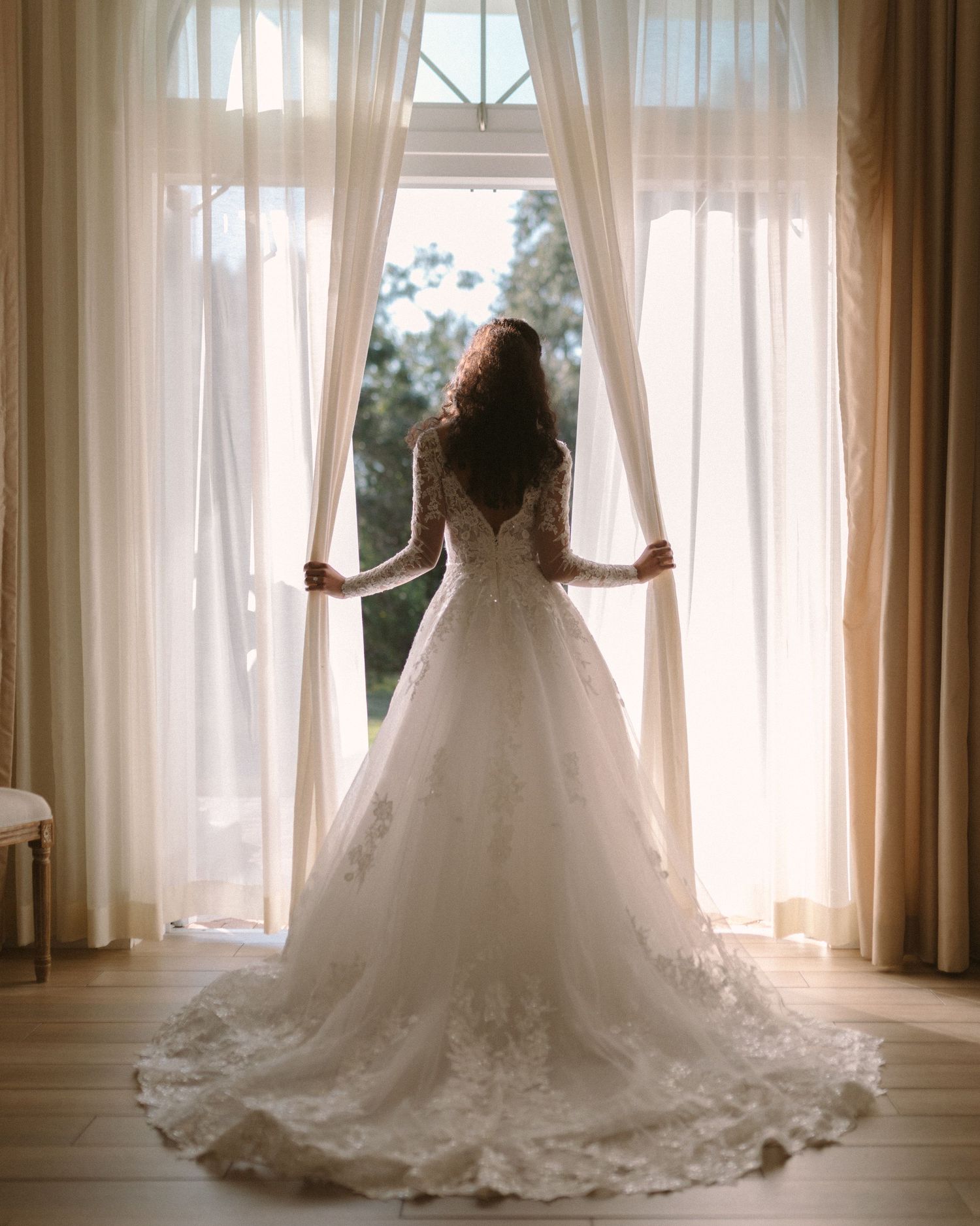 Bride in soft window light, framed through a doorway for an intimate, candid getting-ready moment.