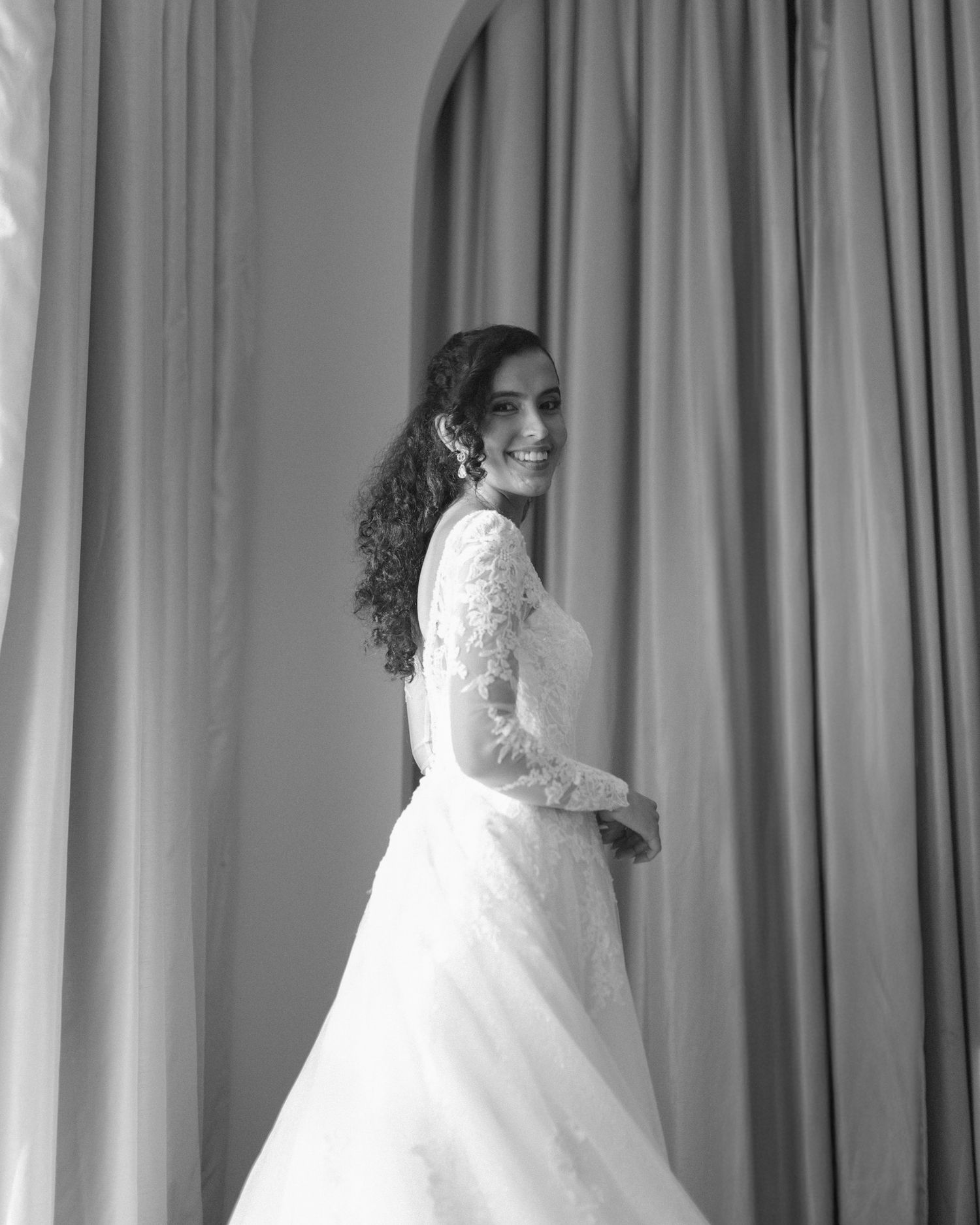 Bride in soft window light, framed through a doorway for an intimate, candid getting-ready moment.