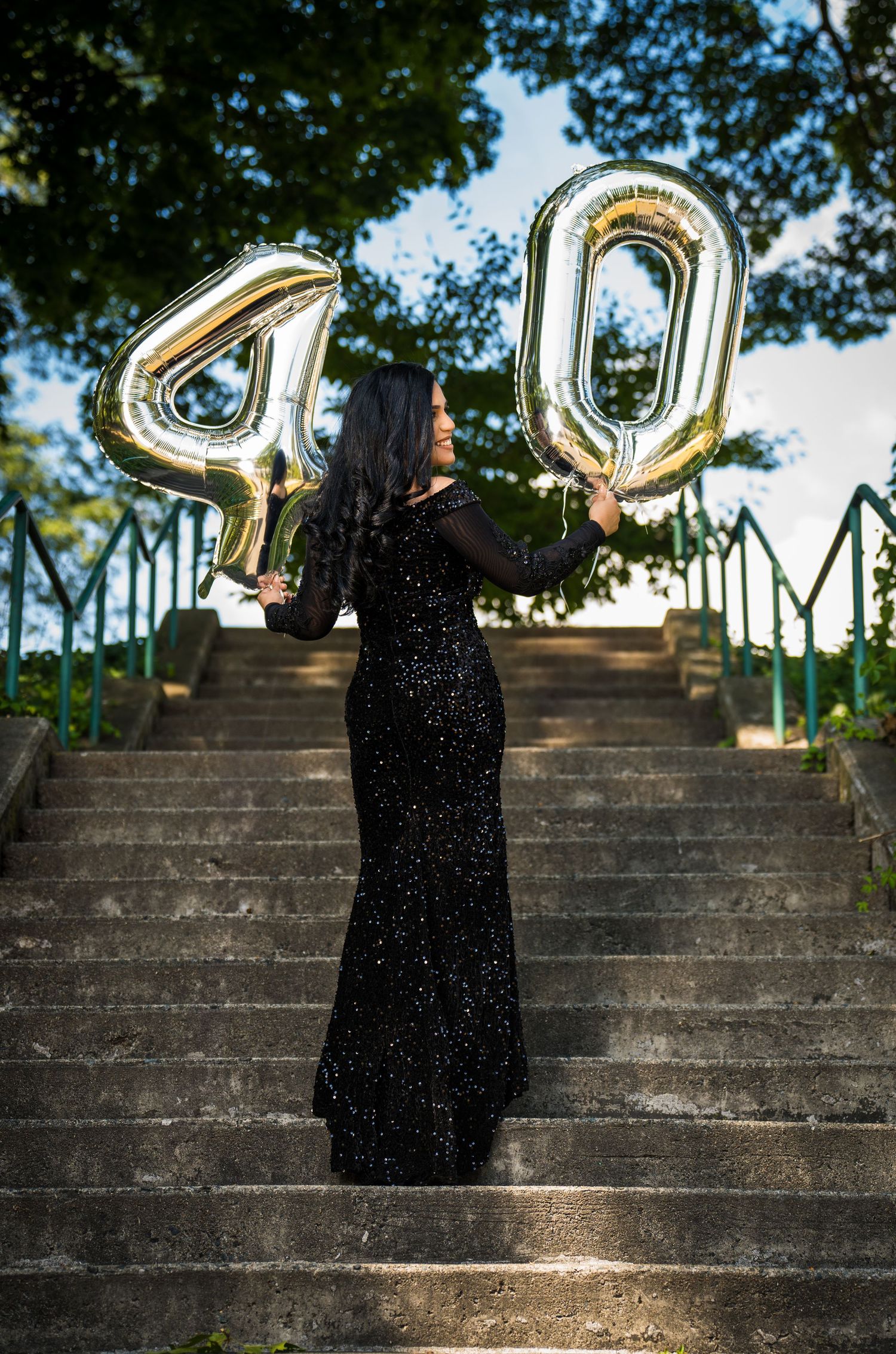 A figure in a sparkly black formal dress holds gold number 40 balloons on stone steps outdoors.