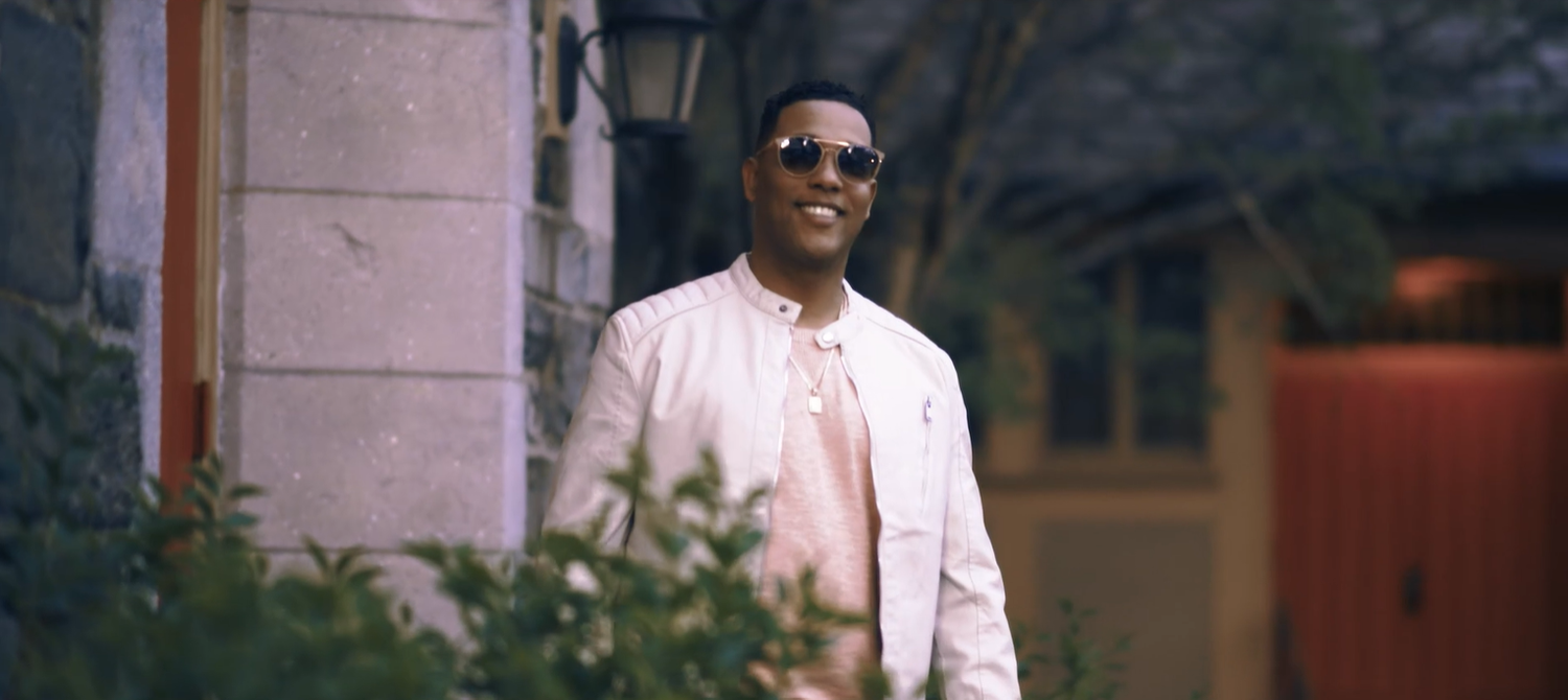 Someone wearing sunglasses and a light button-up shirt stands outside against a blurred background in cinematic lighting.