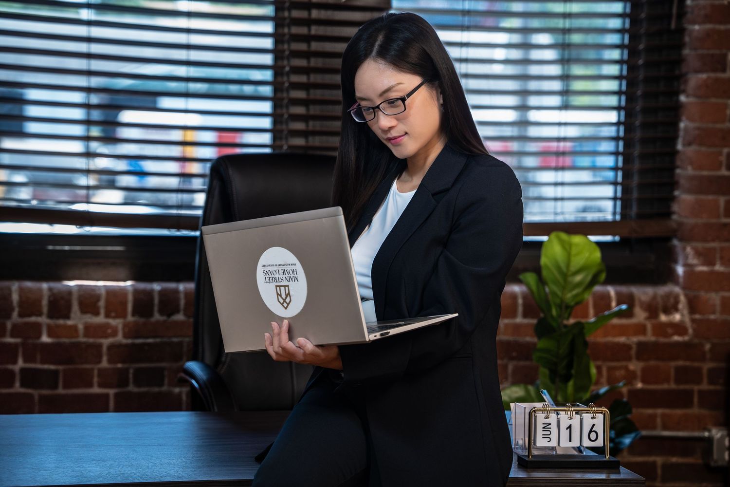 A professional in a business suit works on a laptop in a modern office with exposed brick walls and natural lighting.
