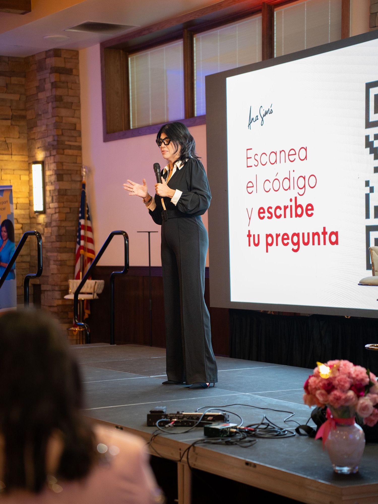 Speaker in black attire presents on stage with a red and white presentation screen showing Spanish text in background.