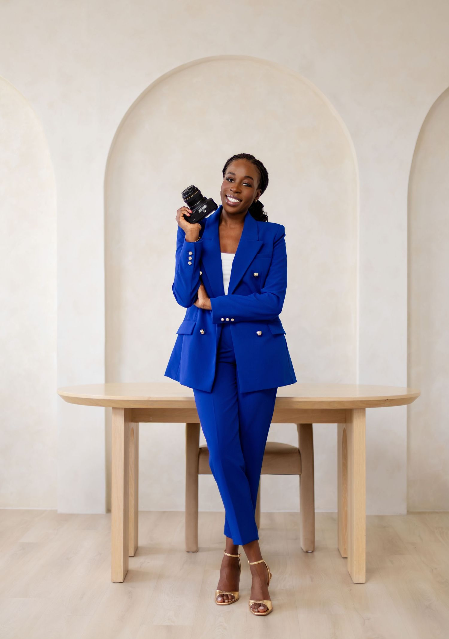 A woman in a royal blue suit holds a camera while leaning on a wooden table in an elegant arched room.