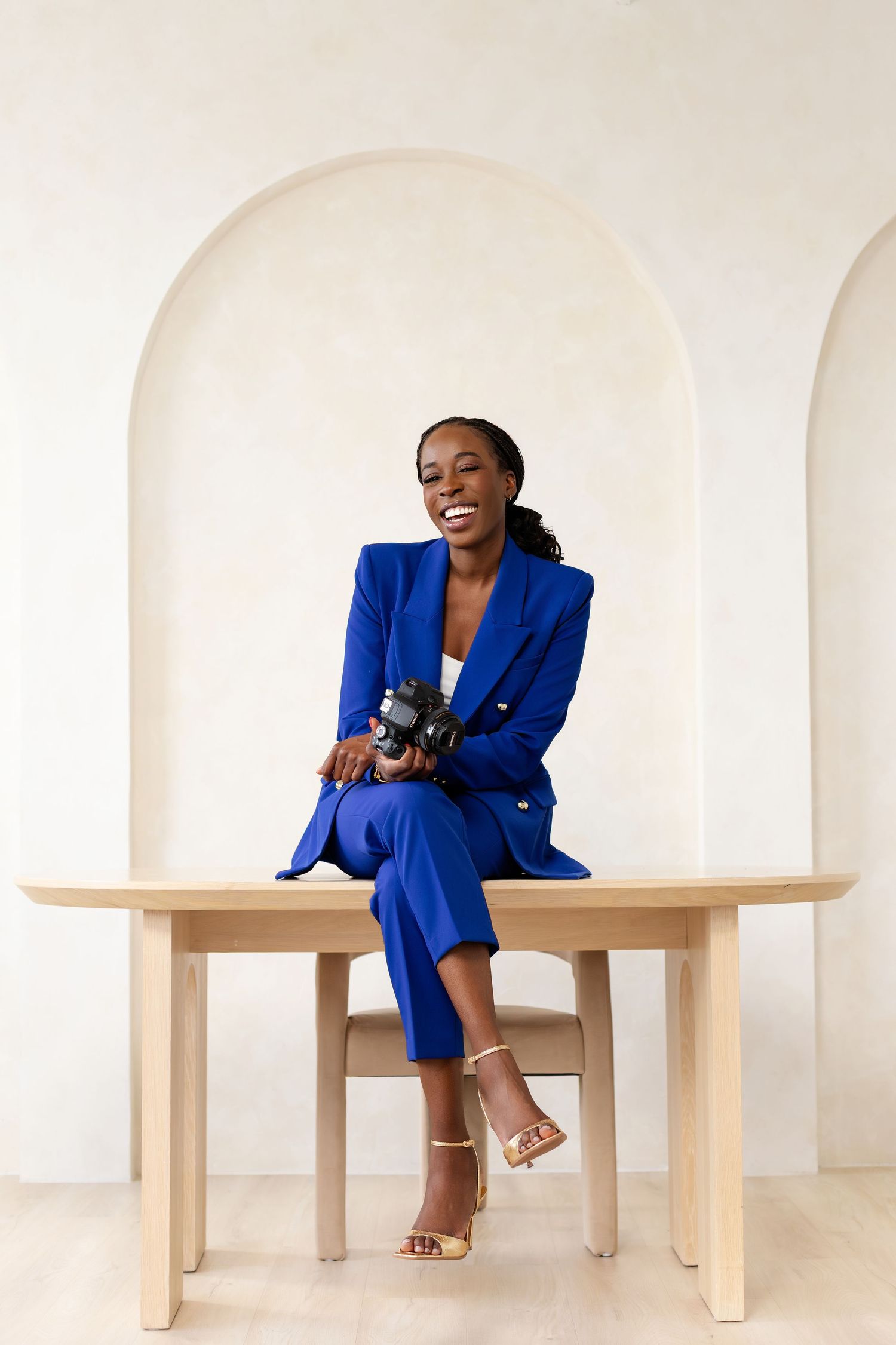 A smiling woman in a royal blue suit sits on a wooden table holding a camera, posed against an arched white wall.