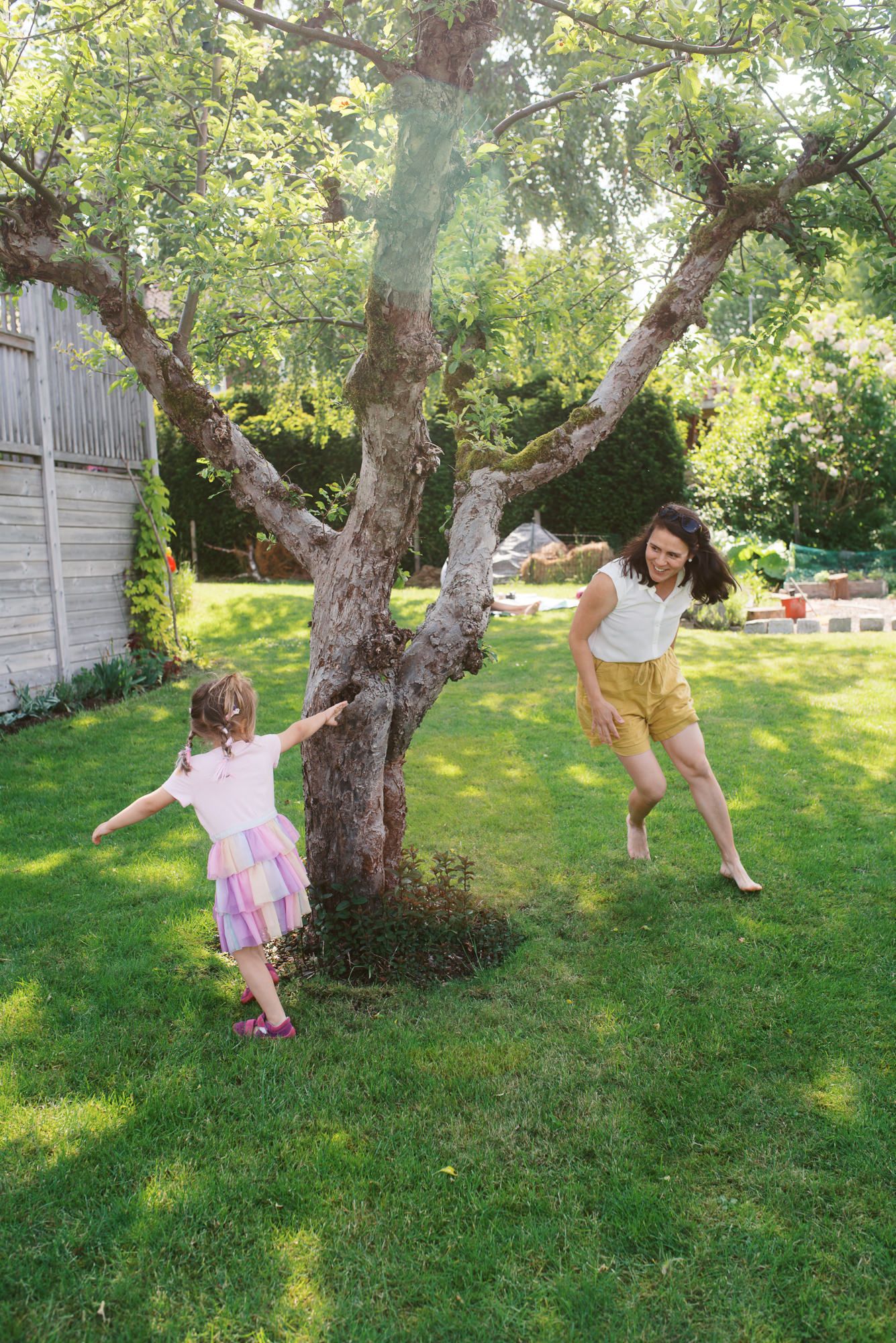Children play and swing from branches of an old apple tree in a sunny backyard garden on a summer afternoon.