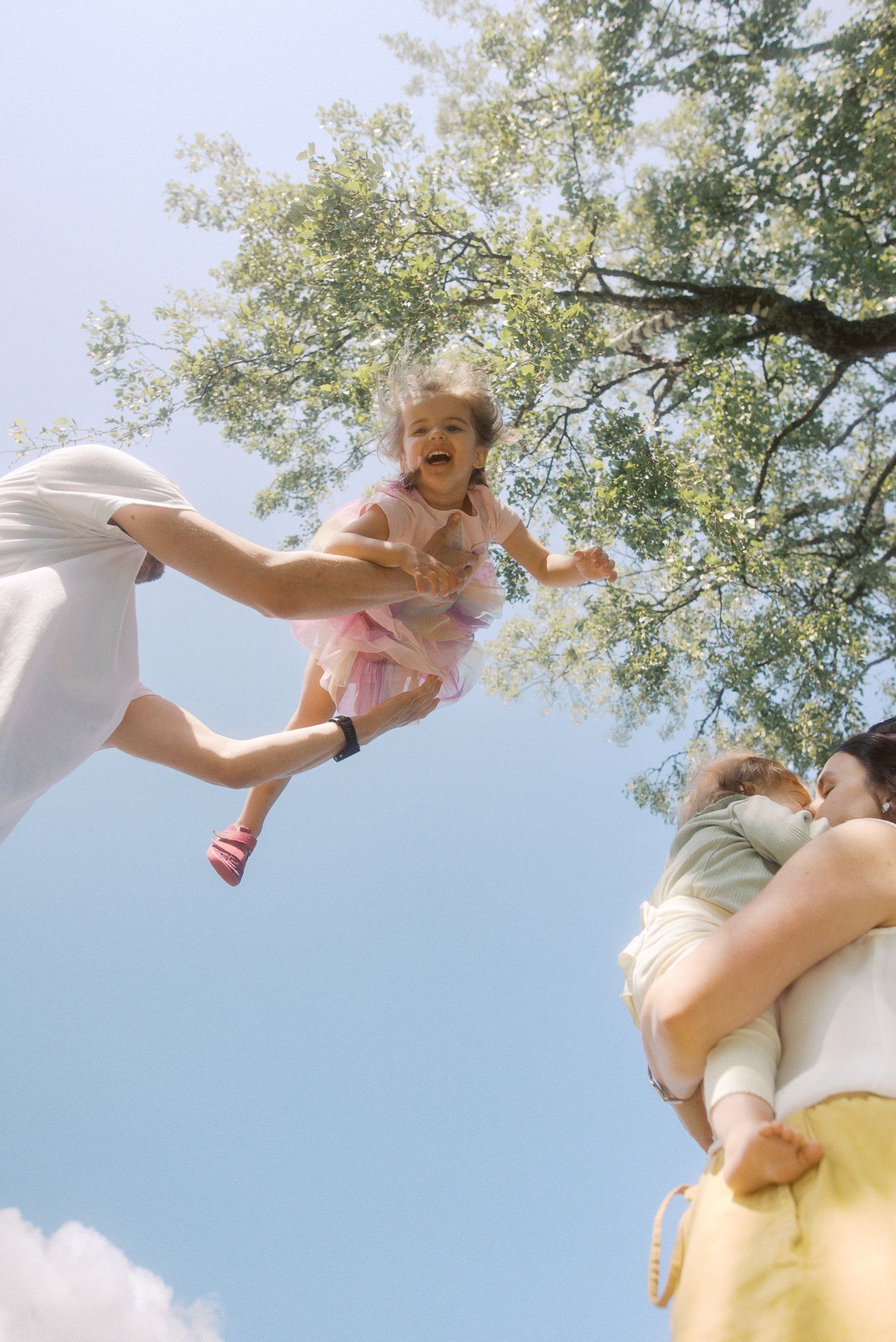 A parent lifts a small child joyfully into the bright blue sky under a leafy tree canopy.