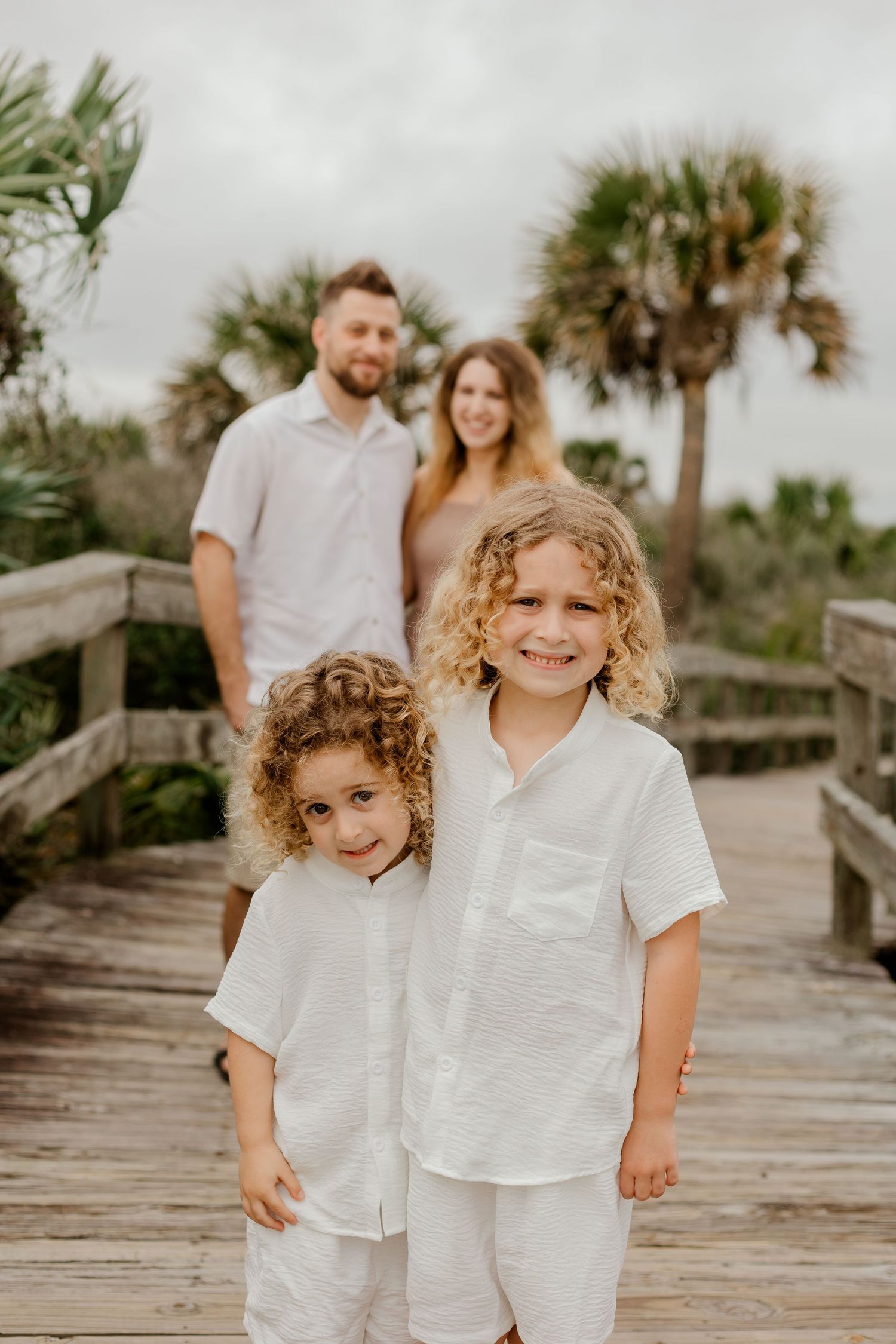 Two curly-haired brothers in white outfits smiling on a boardwalk with parents standing behind them.