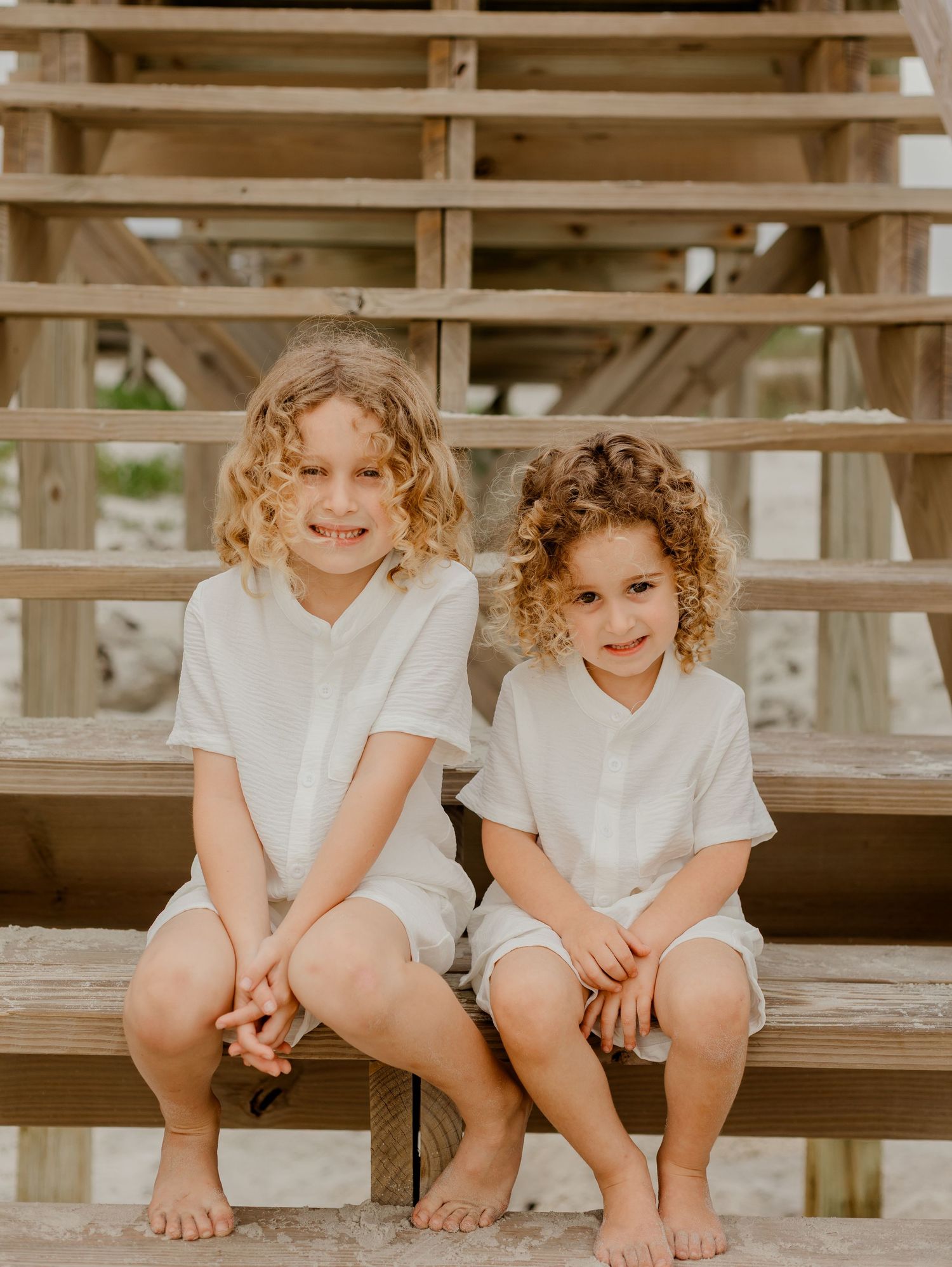 Two curly-haired boys in matching white outfits sitting together on wooden beach stairs, smiling.