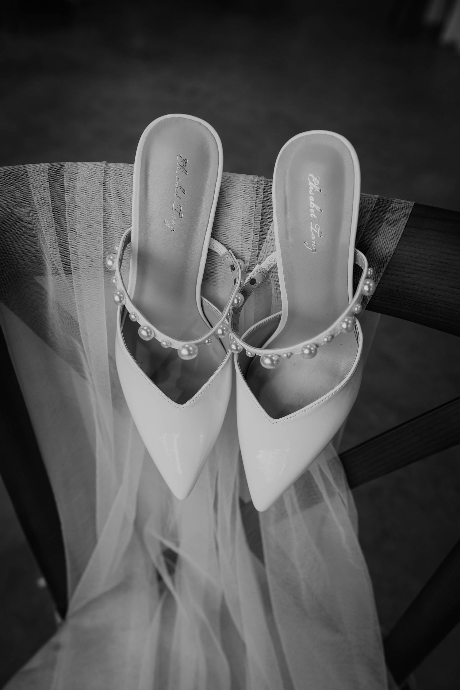 Elegant white bridal heels with pearl embellishments resting on a tulle veil, captured in black and white.