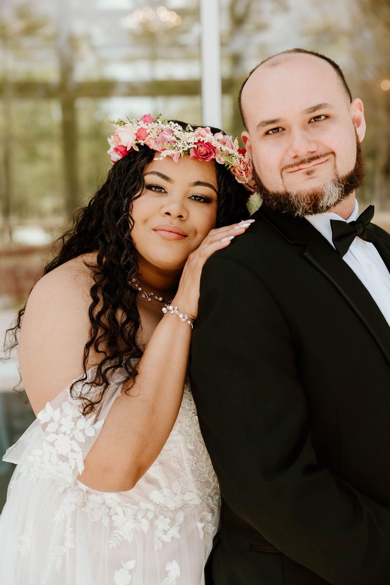 Interracial couple in wedding attire, bride wearing floral crown with pink roses, groom in black tuxedo, posing together.