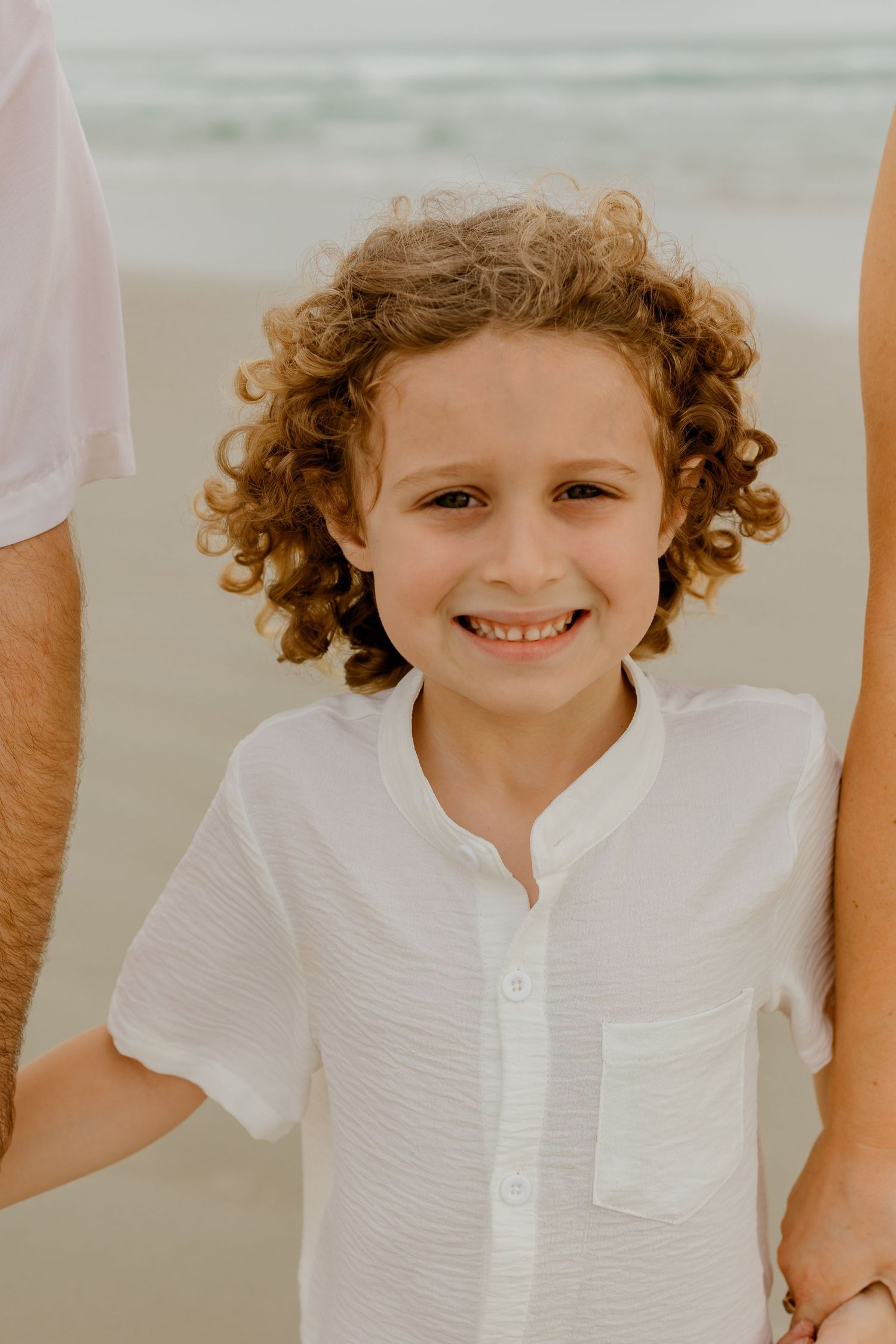 Young curly-haired boy in white shirt smiling at the camera while standing on a sandy beach.