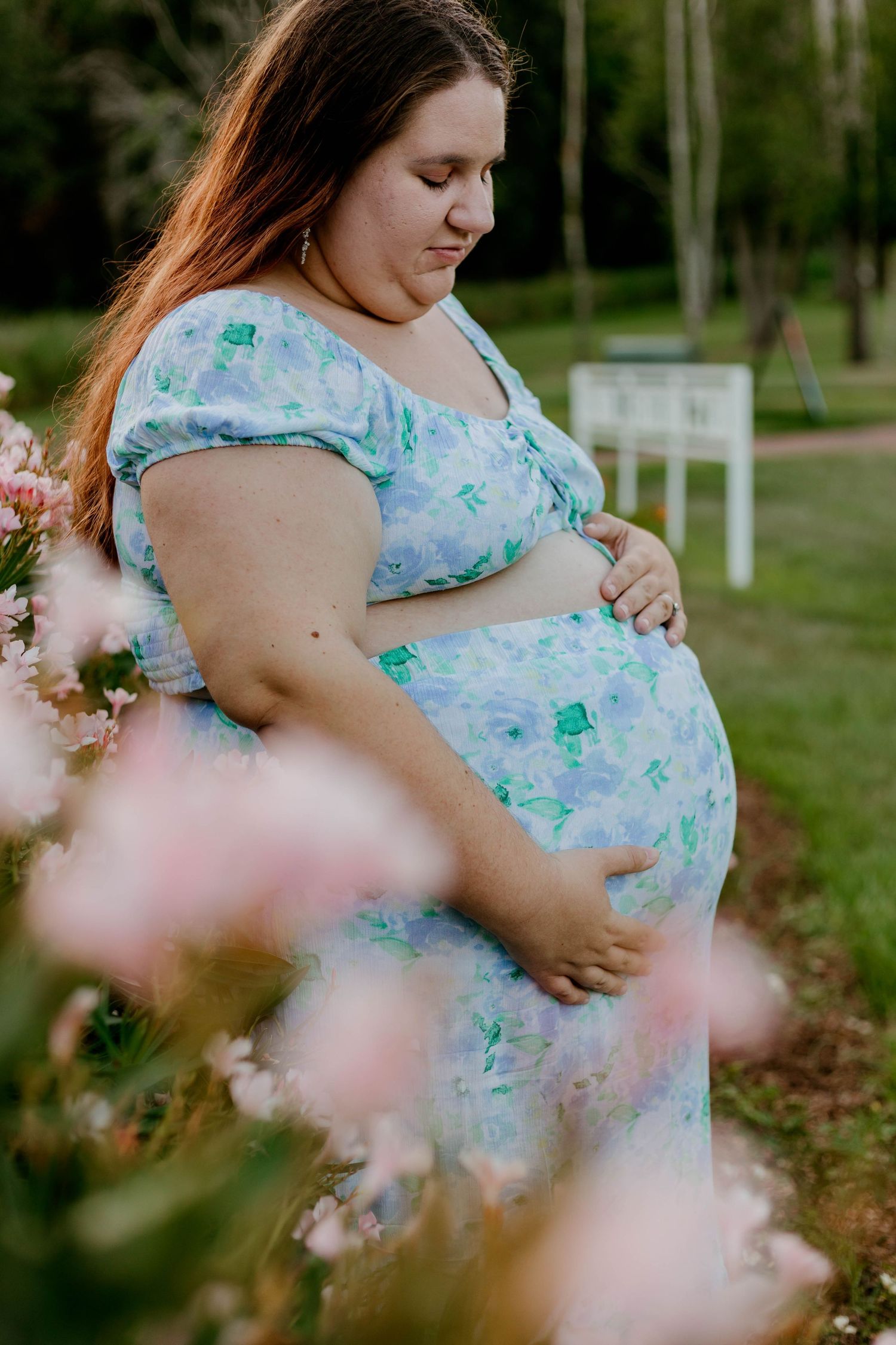 Pregnant woman in floral dress gently holding baby bump surrounded by soft pink flowers outdoors.