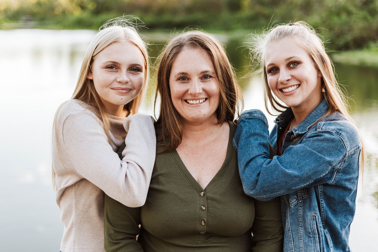 A smiling mother poses outdoors with her two blonde daughters near a peaceful lake.