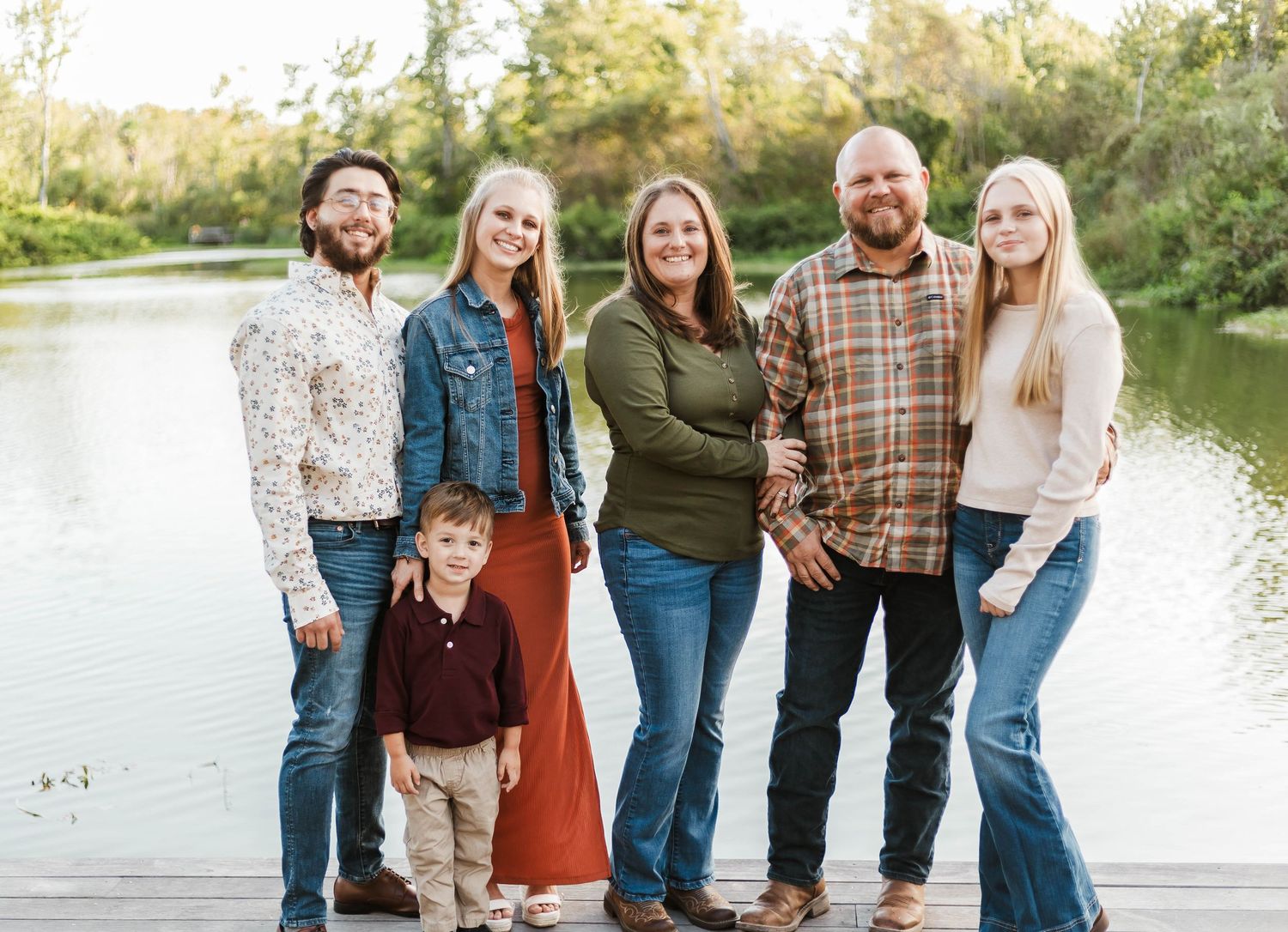 Family of six posing together on a wooden dock by a serene lake surrounded by lush green trees.