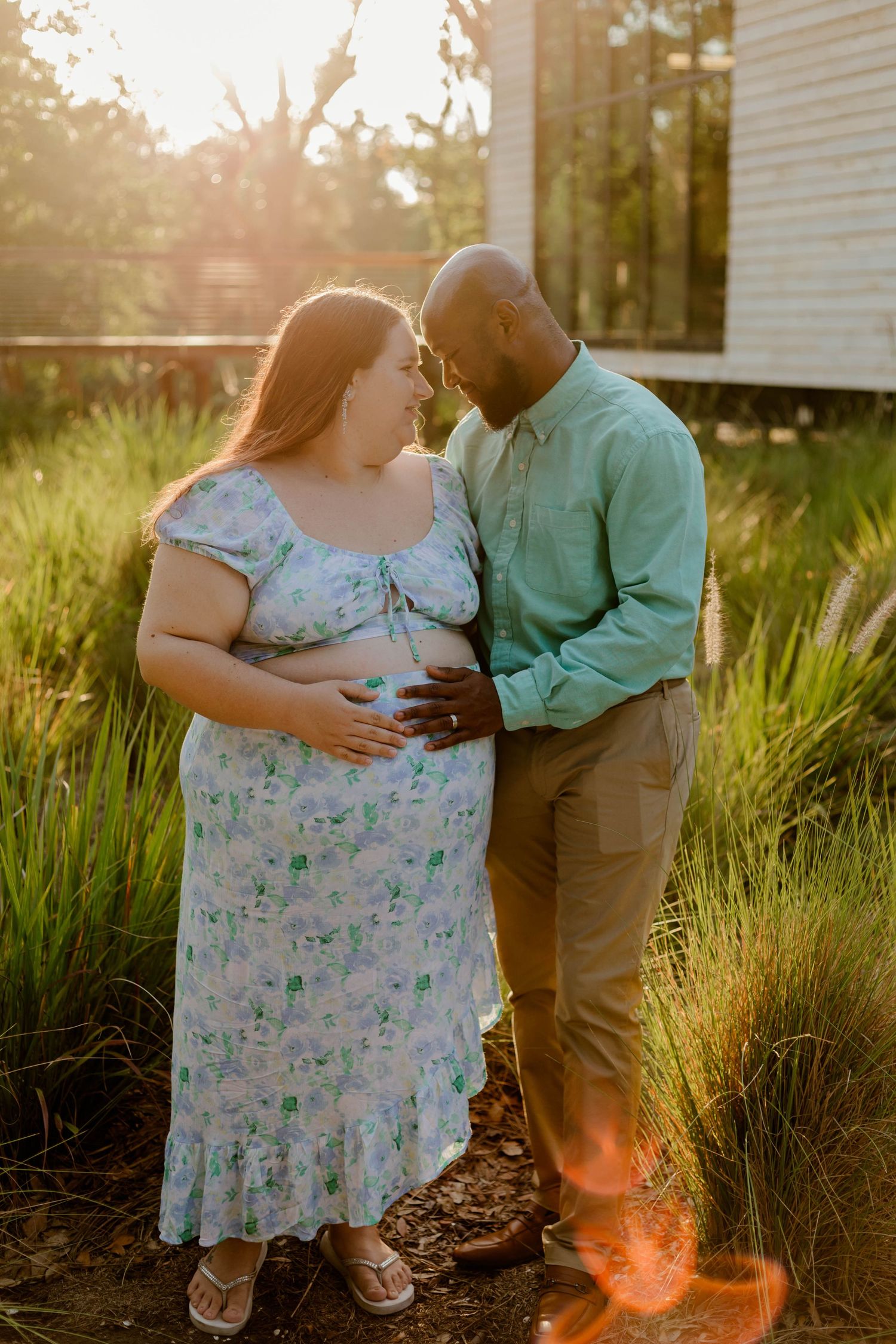 Expectant interracial couple touching foreheads lovingly while cradling baby bump in golden sunlight.