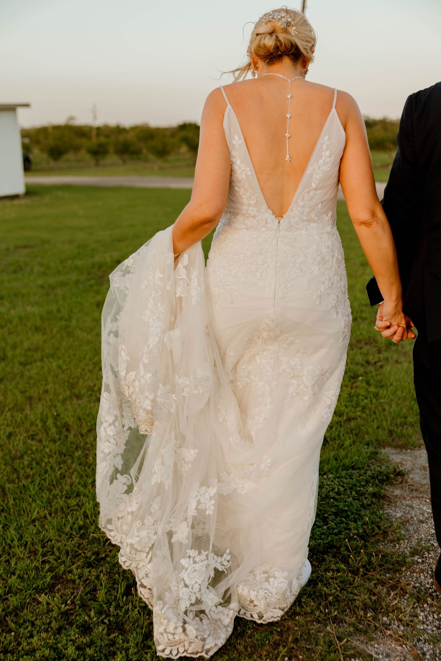 Bride in backless lace wedding gown holding train while walking hand in hand with groom at sunset.