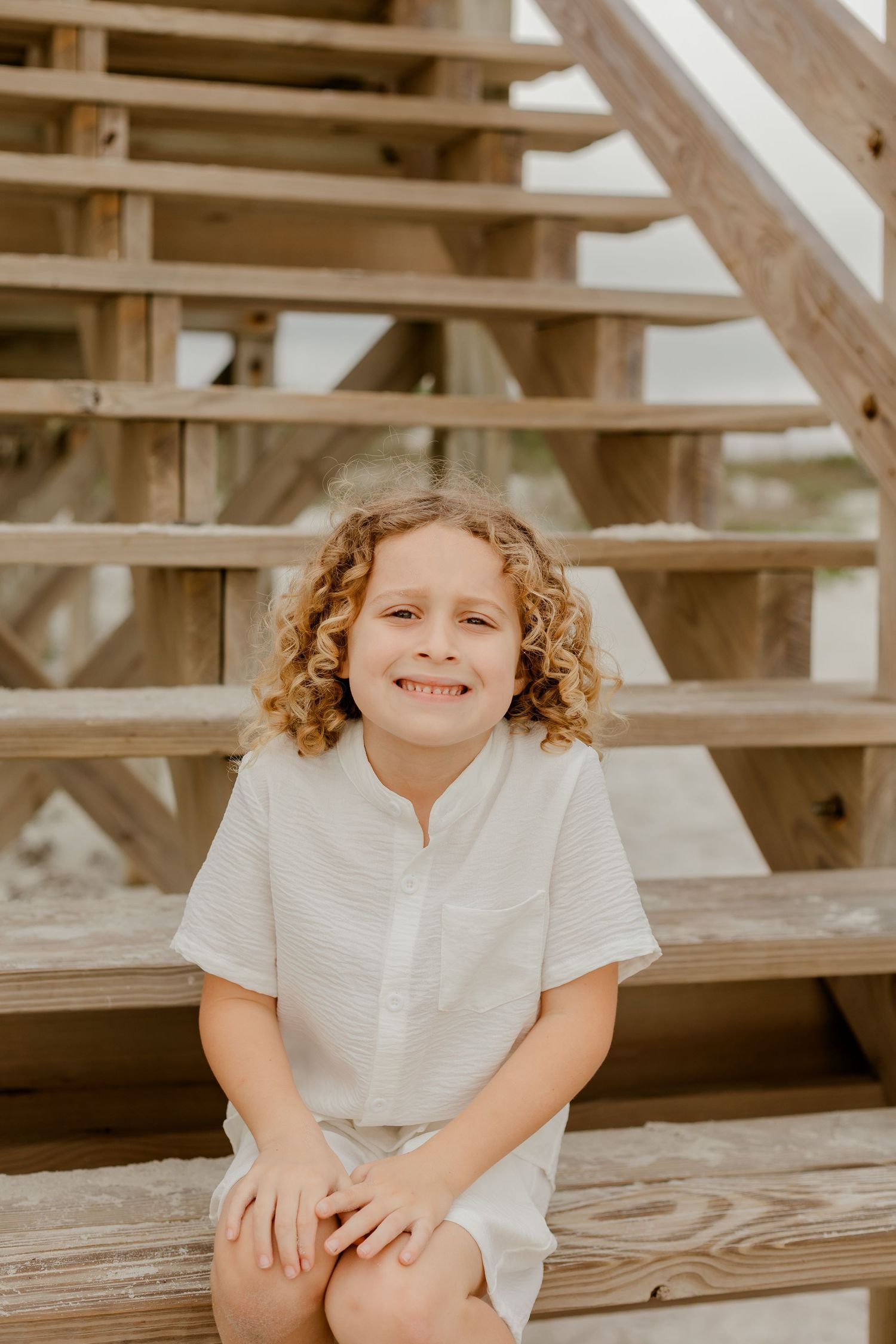 Curly-haired boy in white outfit sitting on wooden beach stairs, smiling at the camera.