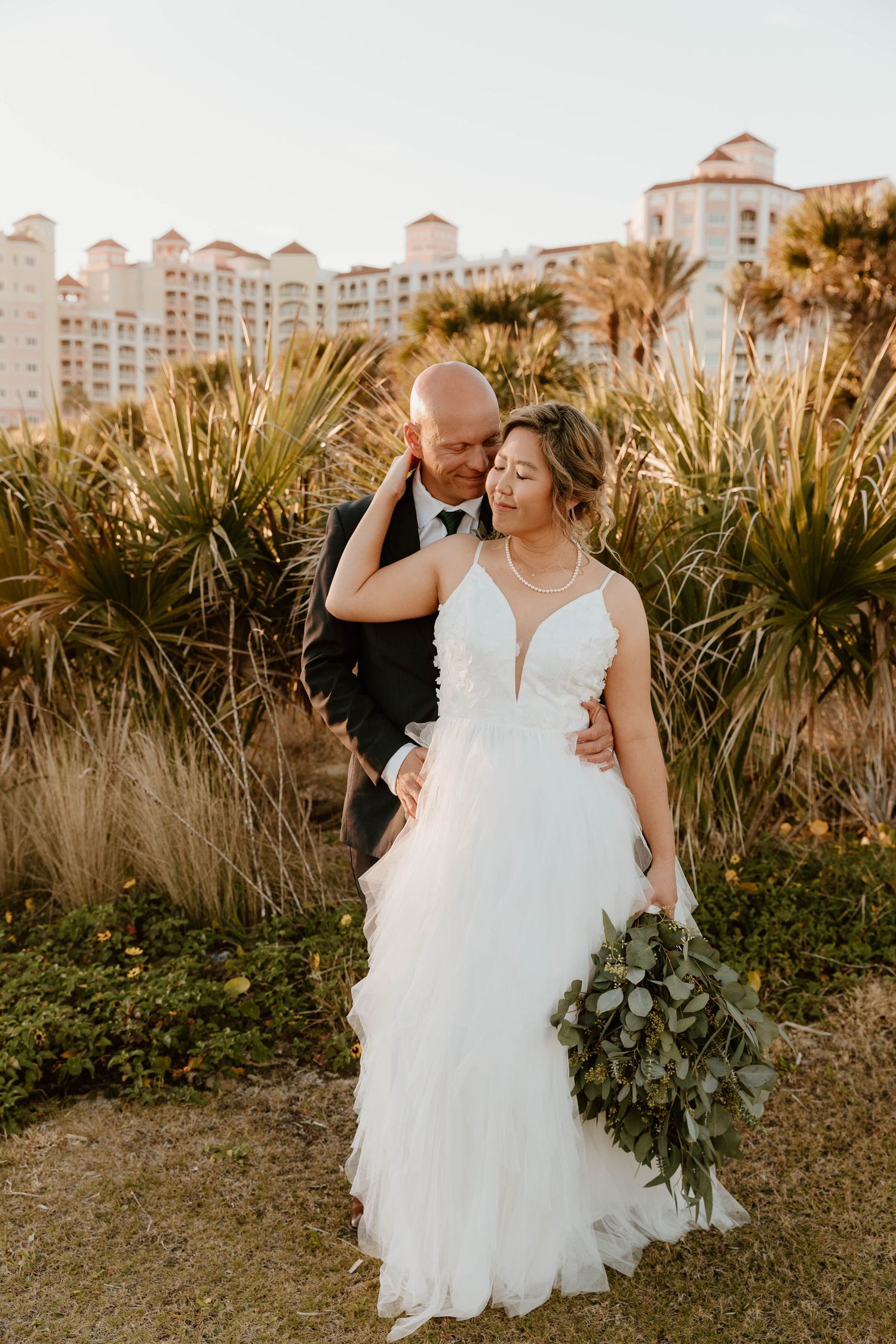 Romantic couple portrait with bride in a plunging tulle gown holding greenery bouquet, palm trees and resort backdrop.