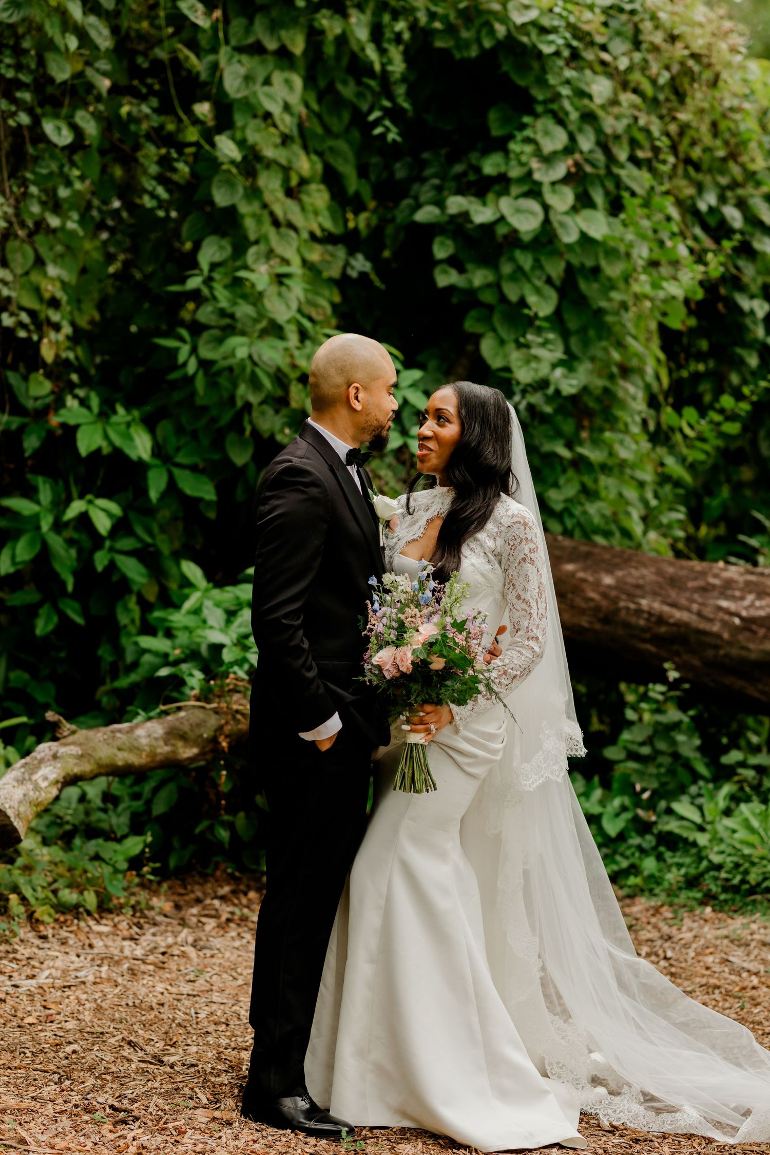 Bride and groom sharing an intimate moment in a lush green forest setting, bride holding a wildflower bouquet.