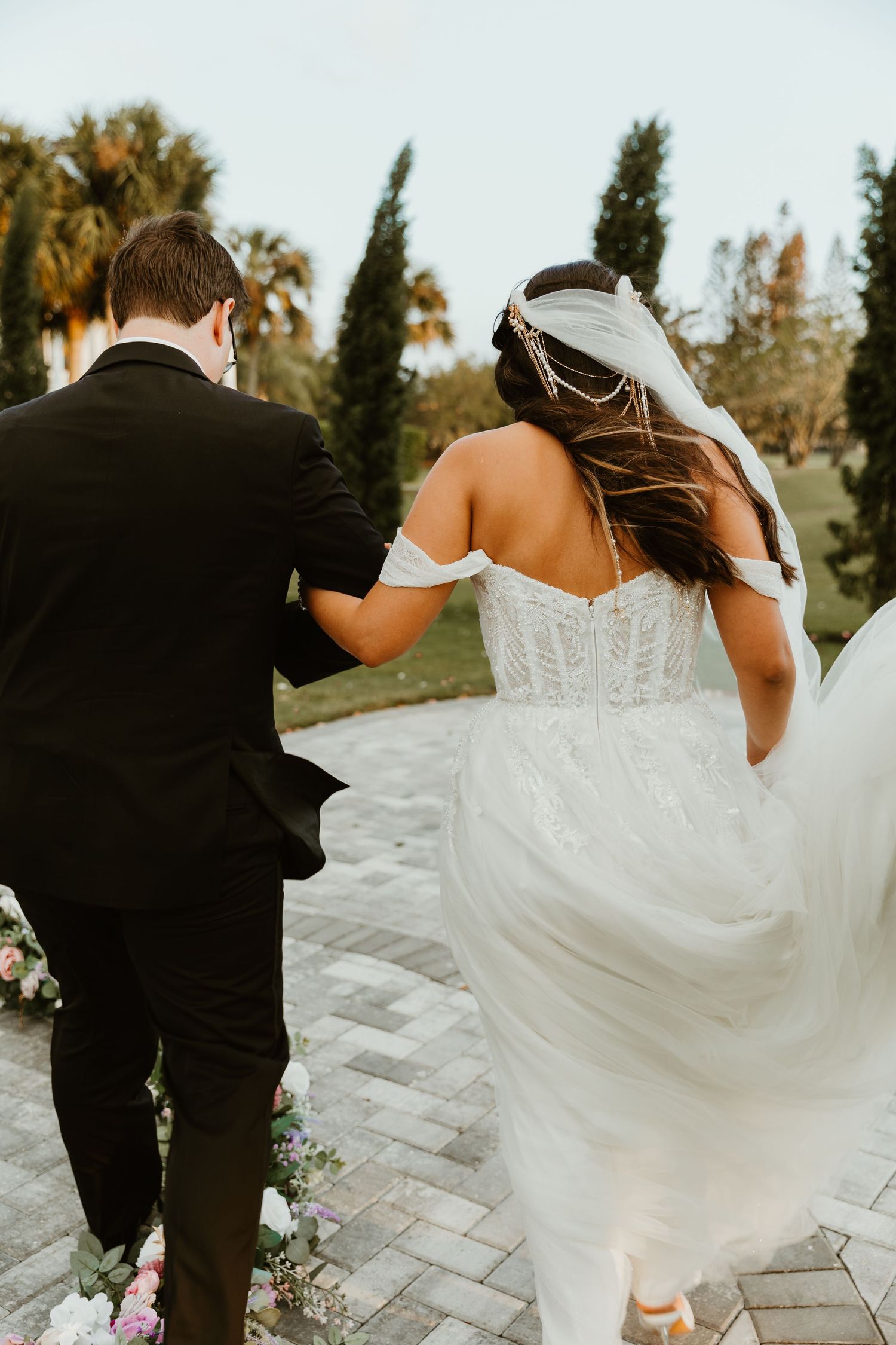 Bride and groom walking away on brick path, bride in off-shoulder white gown with flowing skirt and veil.