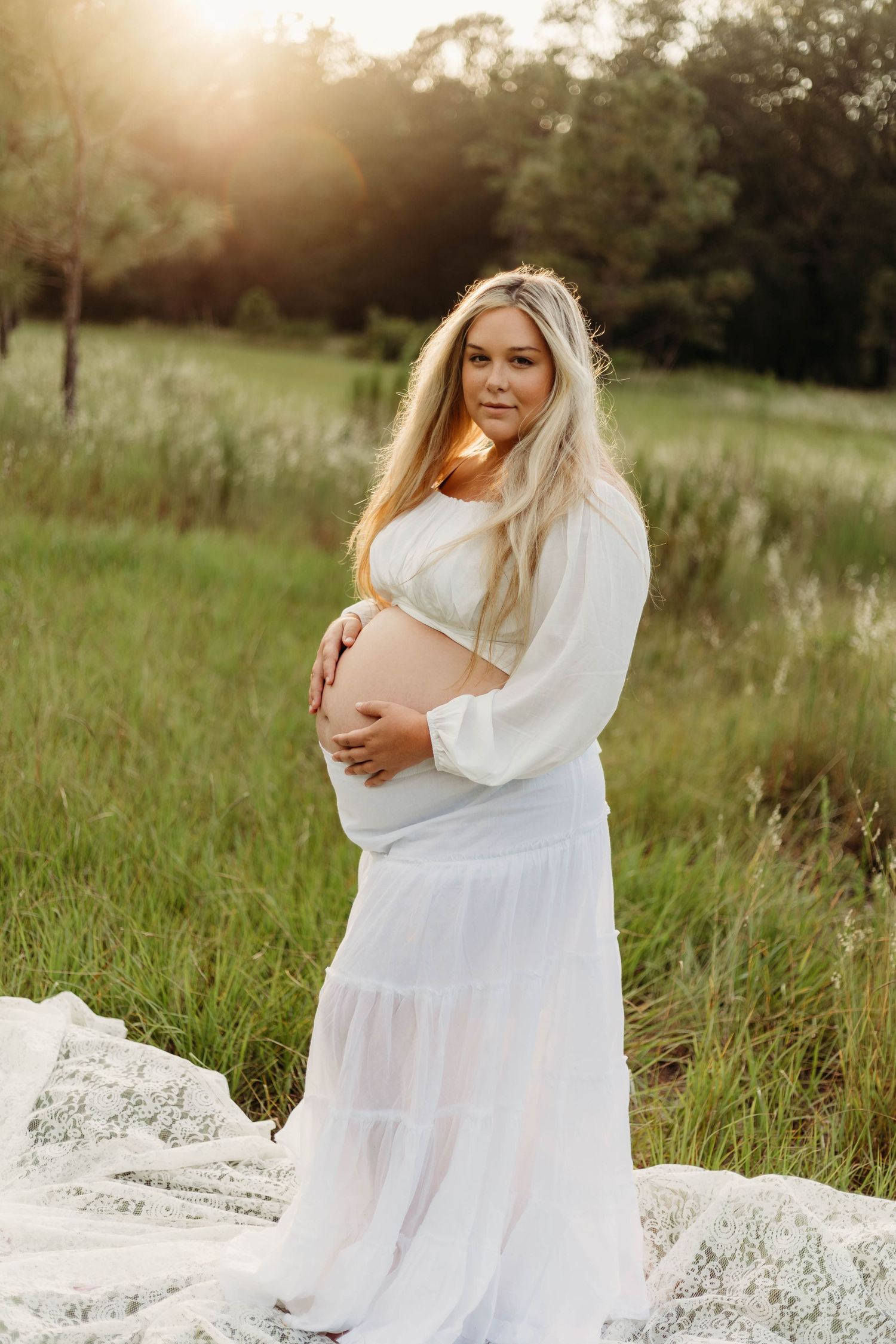 Pregnant woman in white lace dress cradles baby bump while posing in sunlit green field at golden hour.