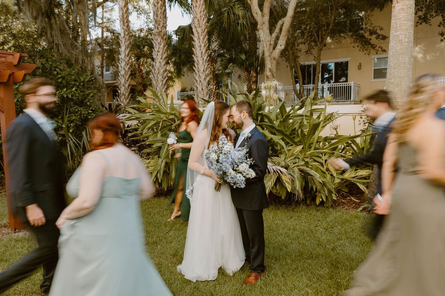 Bride and groom share a romantic moment outdoors surrounded by tropical palms and blurred wedding guests.