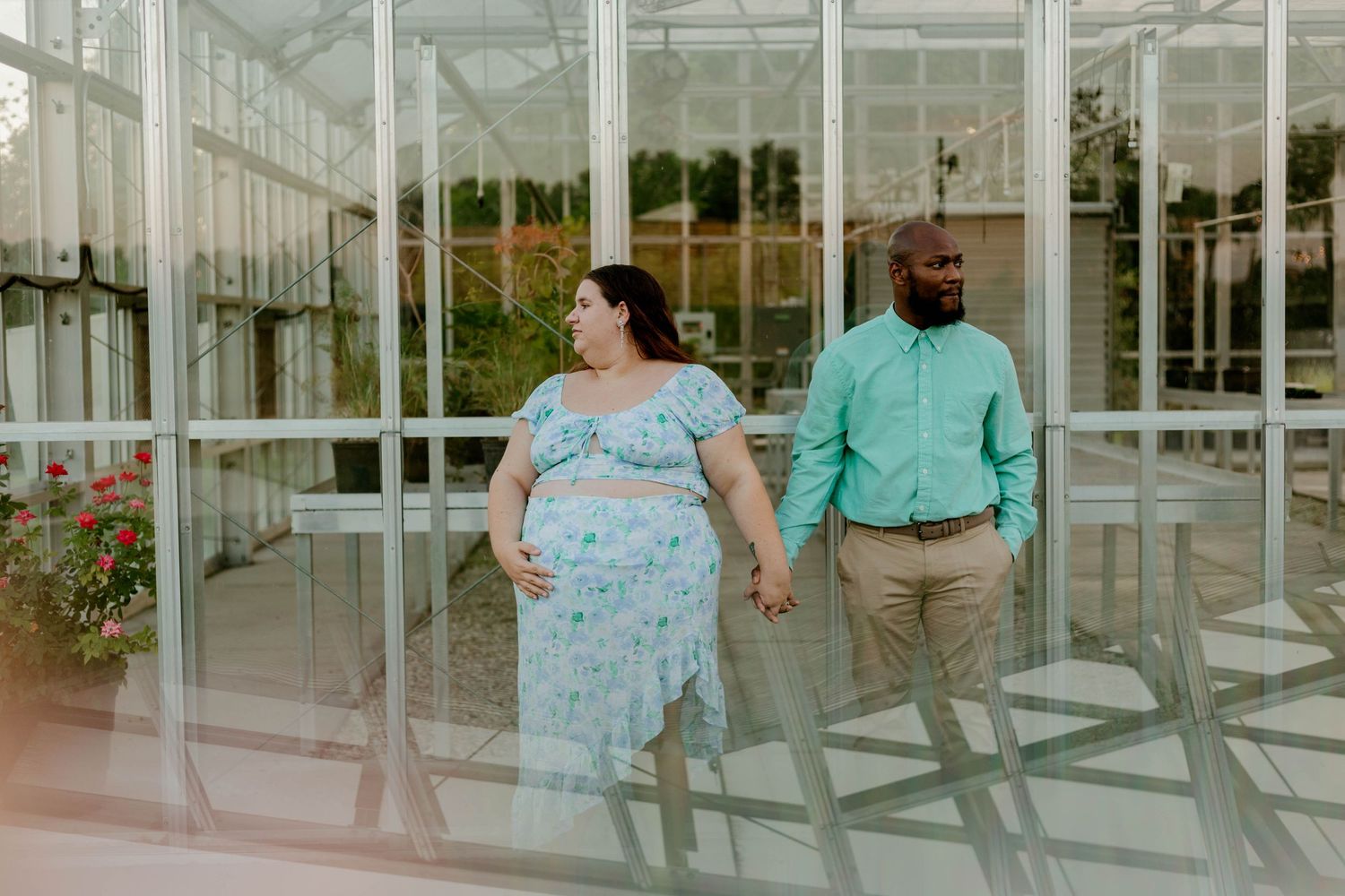 Expectant interracial couple holding hands outside a greenhouse, woman in floral maternity dress.