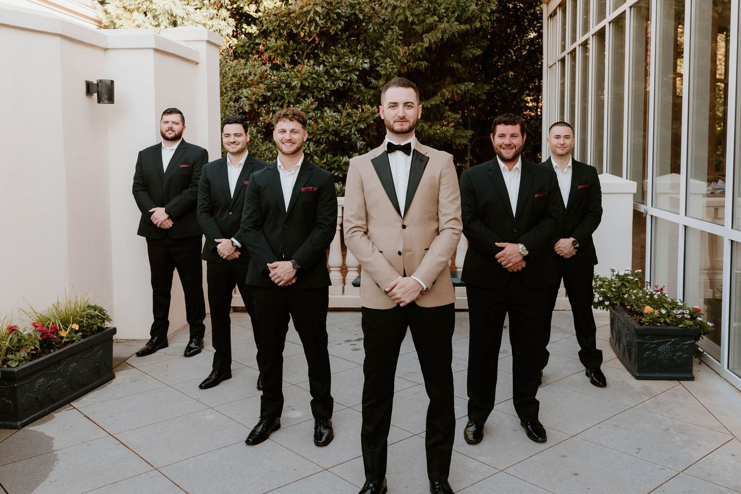 Groom in tan tuxedo stands confidently before four groomsmen in black suits on a courtyard patio.