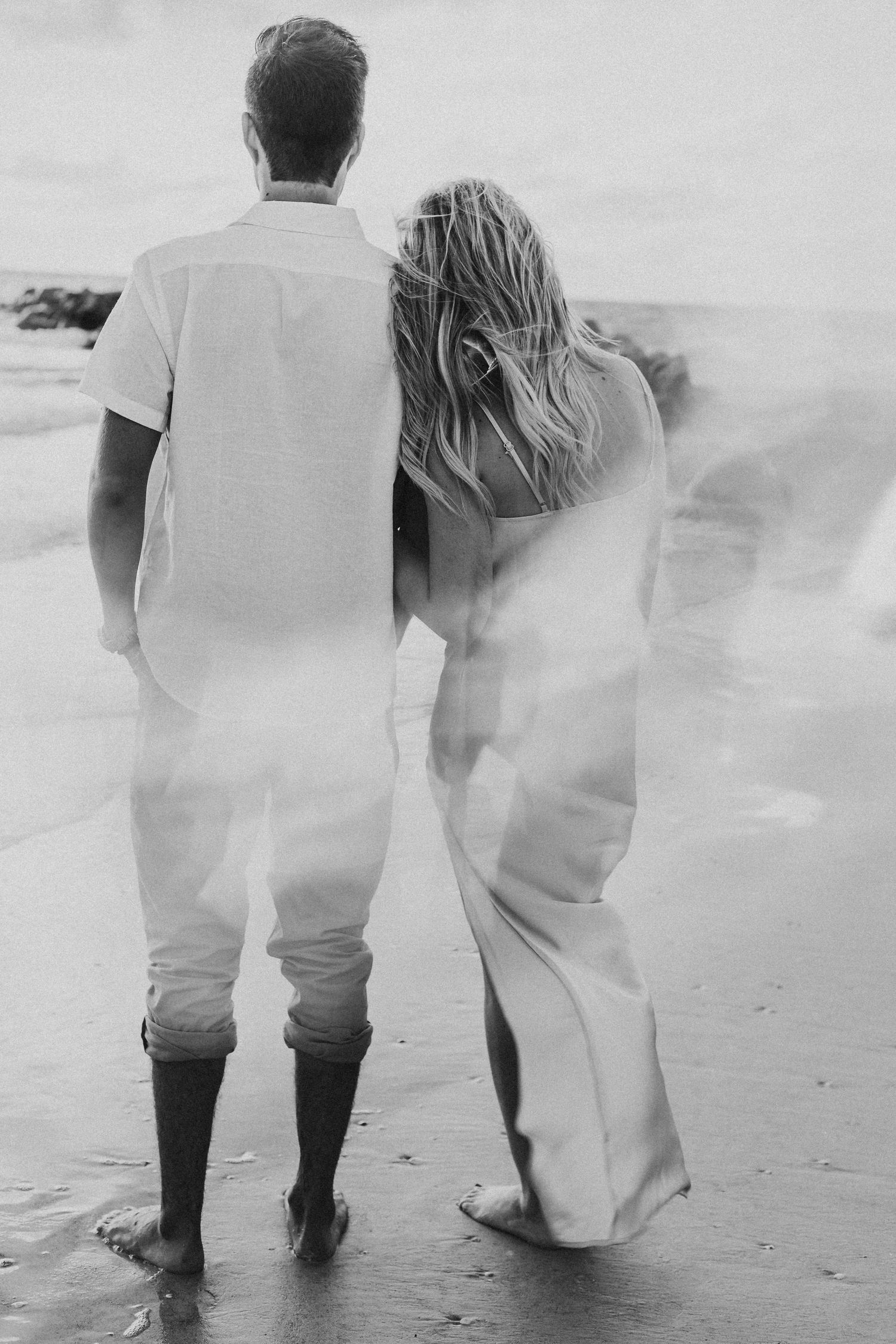 Black and white photo of couple standing together on windy beach, woman in flowing white dress.