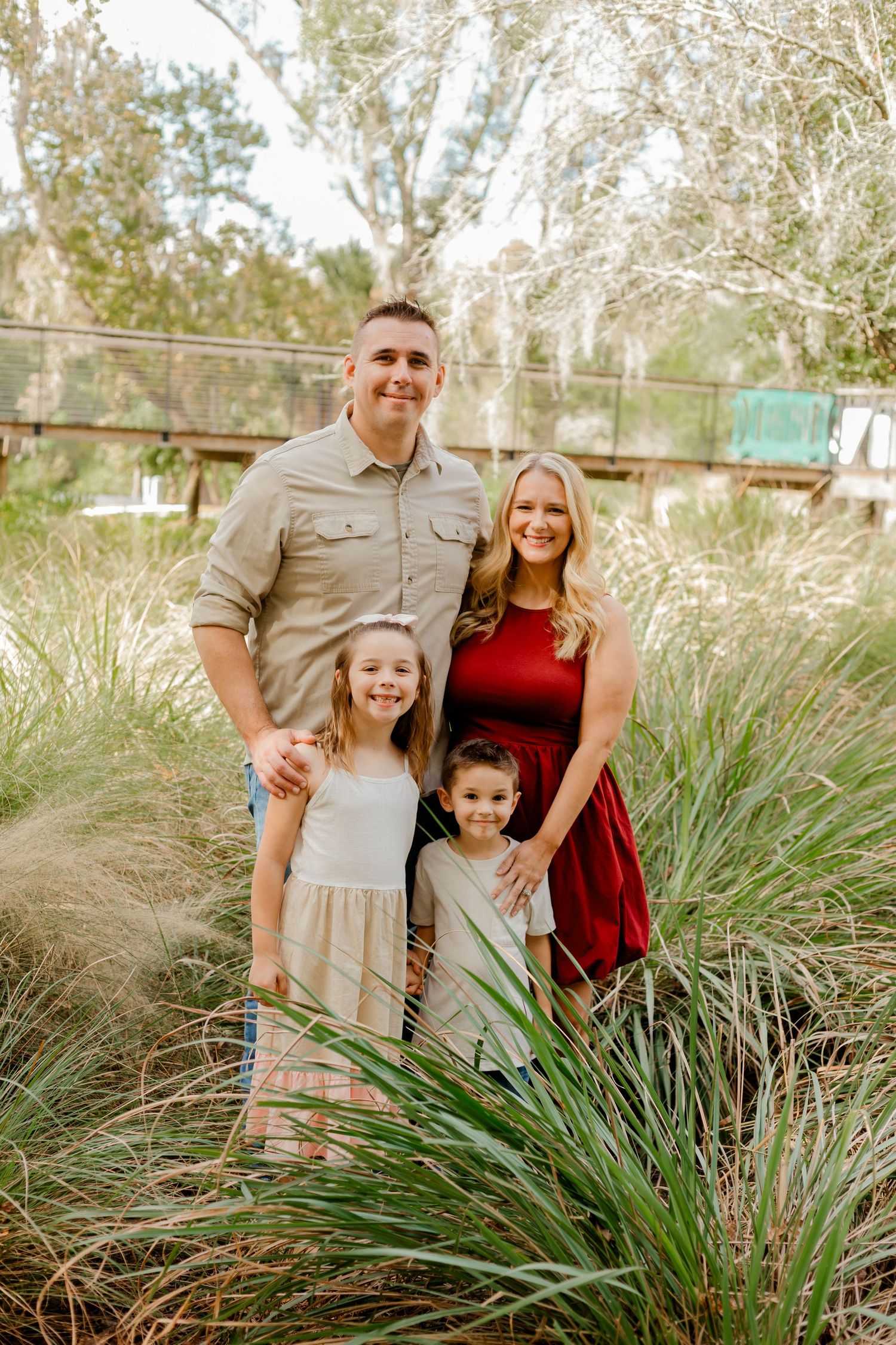 Family of four posing among lush greenery, parents smiling with two young children in a sunny outdoor setting.