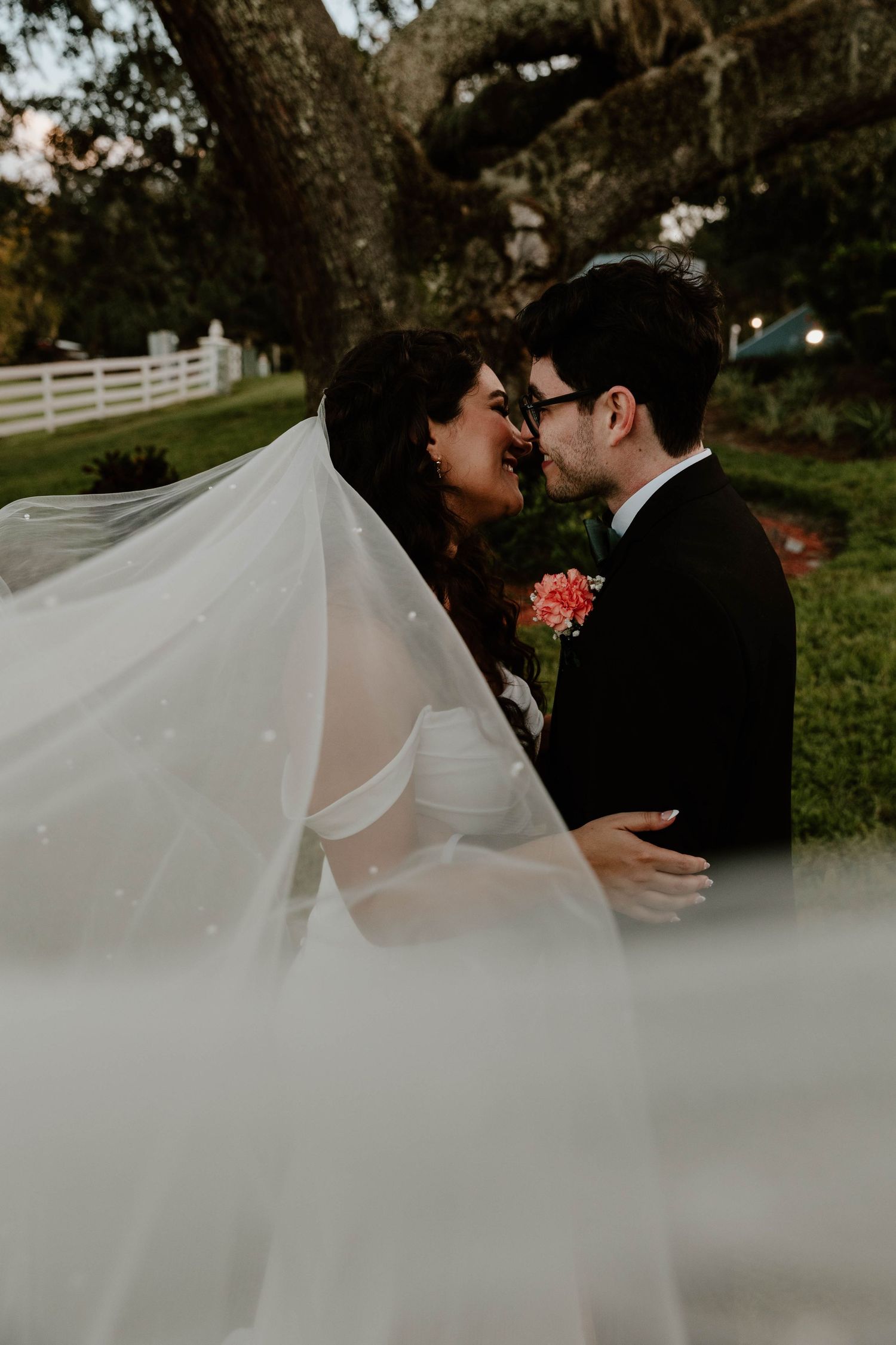 A bride and groom share a romantic moment under a large oak tree, with her flowing veil billowing in the foreground.
