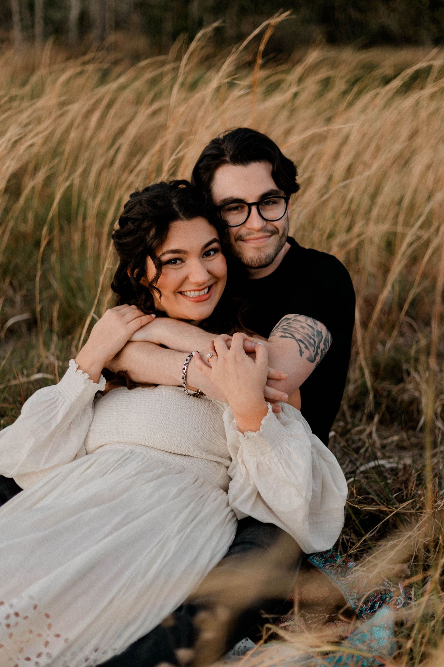 Expecting couple sitting in tall golden grass, man embracing pregnant woman in white dress.