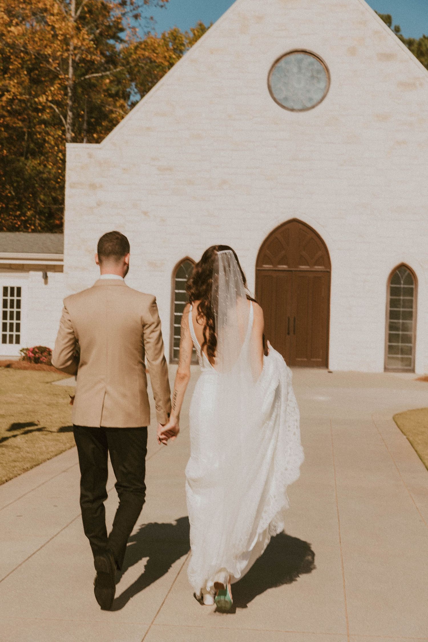 Newlywed couple holding hands walking toward a white brick chapel with arched wooden doors on a sunny day.