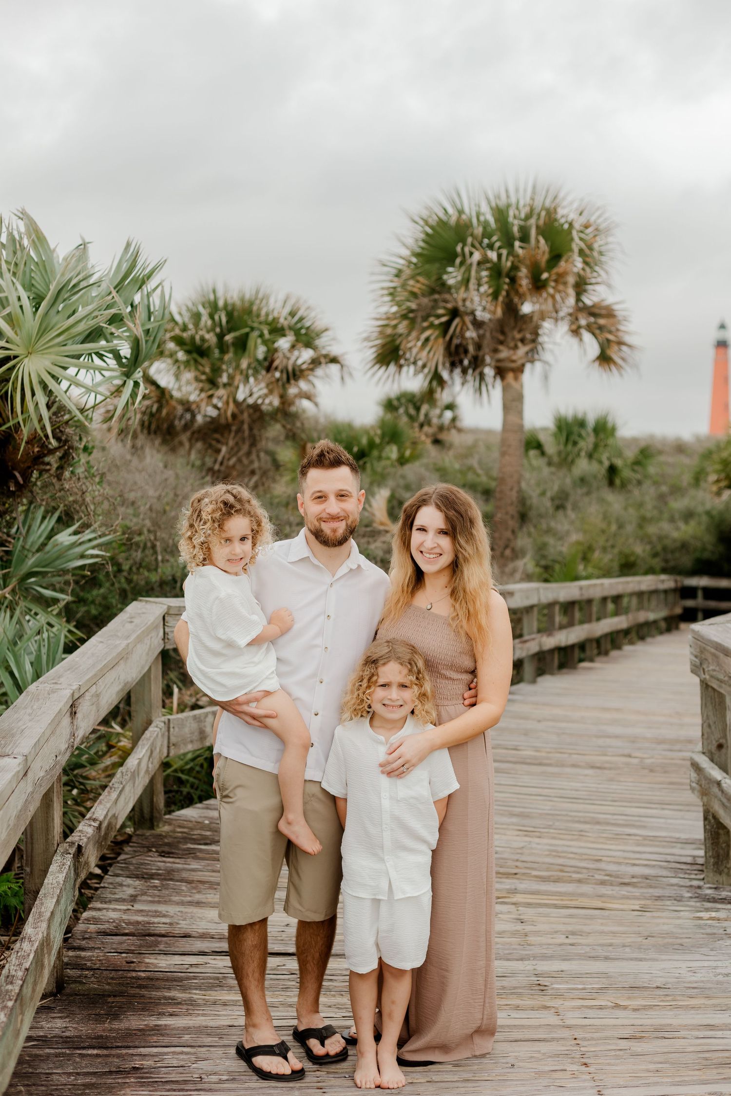 Family of four posing on a wooden boardwalk with palm trees and a lighthouse in the background.