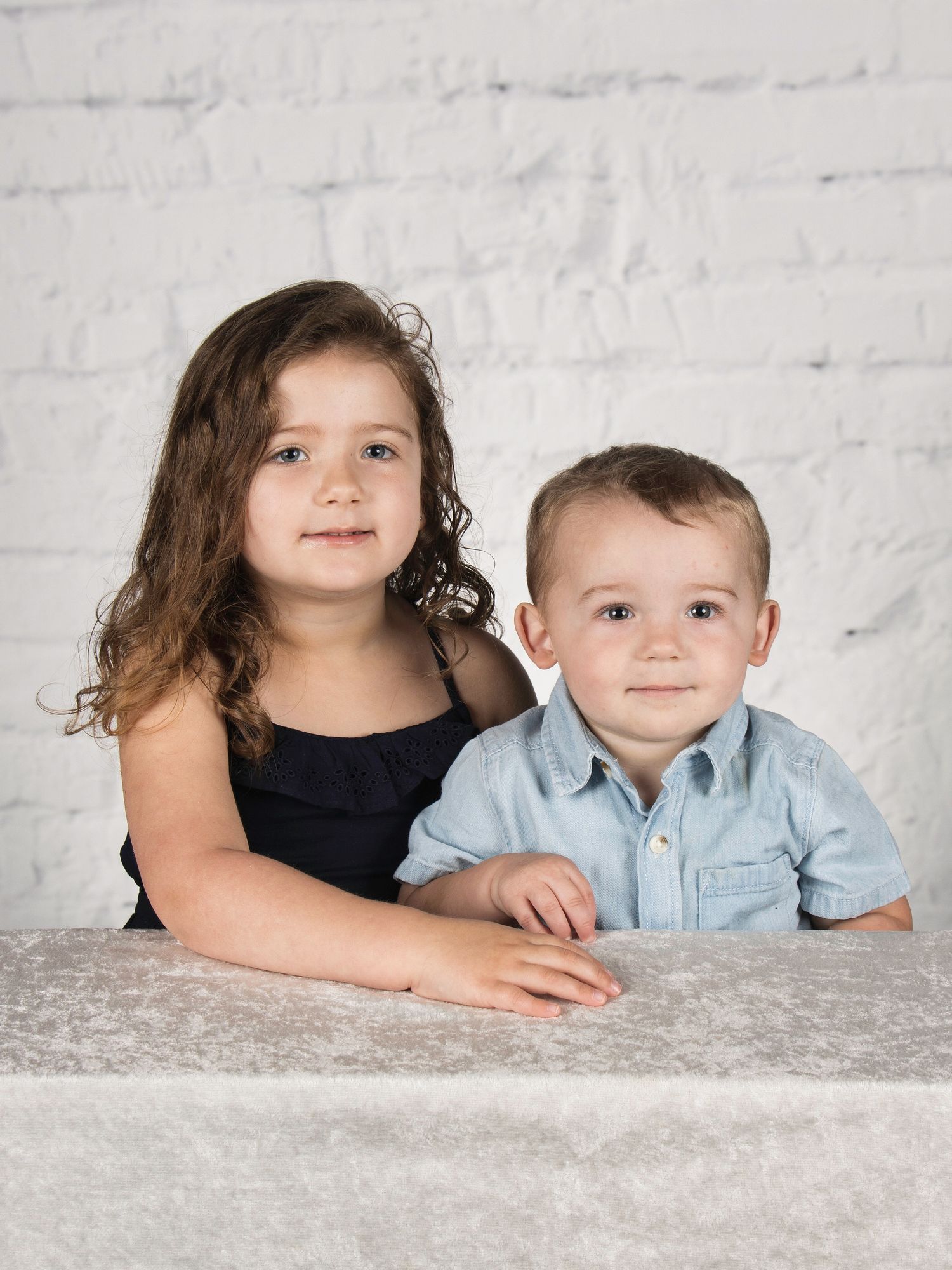 Two young siblings, a curly-haired girl in black and a toddler boy in blue, pose at a table.