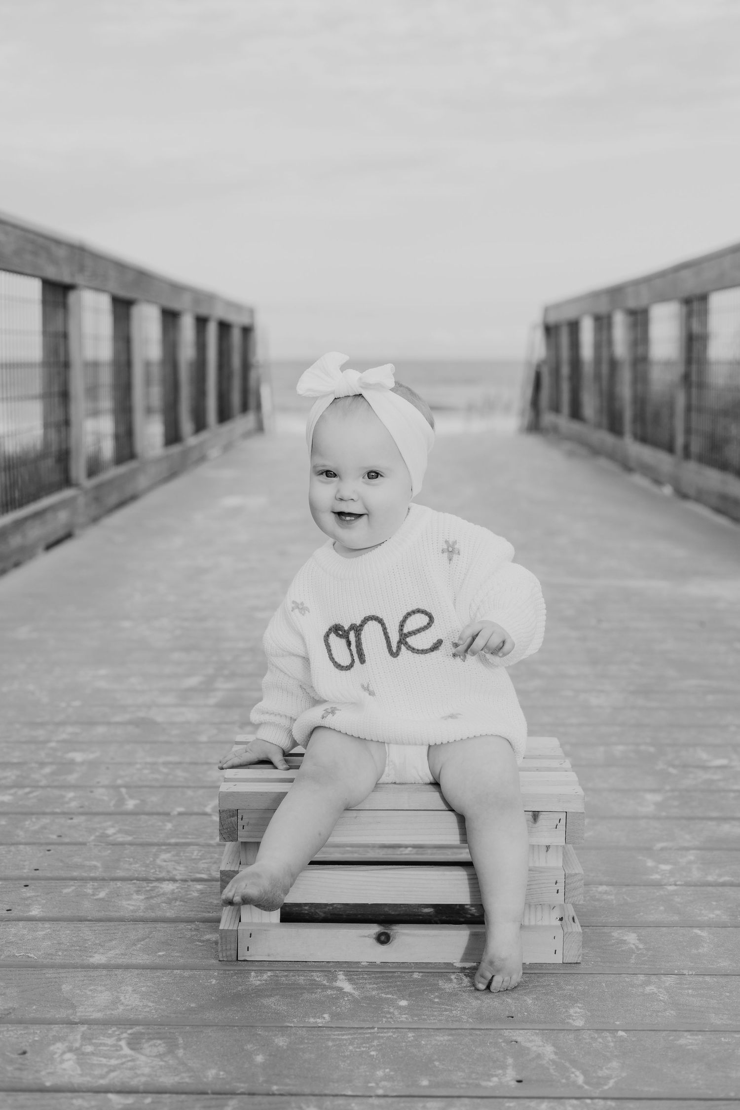 Black and white photo of a smiling baby wearing a bow headband sitting on a crate on a boardwalk.