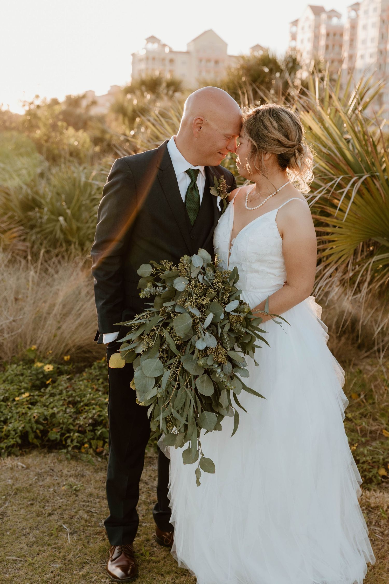 Bald groom in dark suit and bride in white gown touching foreheads in a sunlit garden setting.