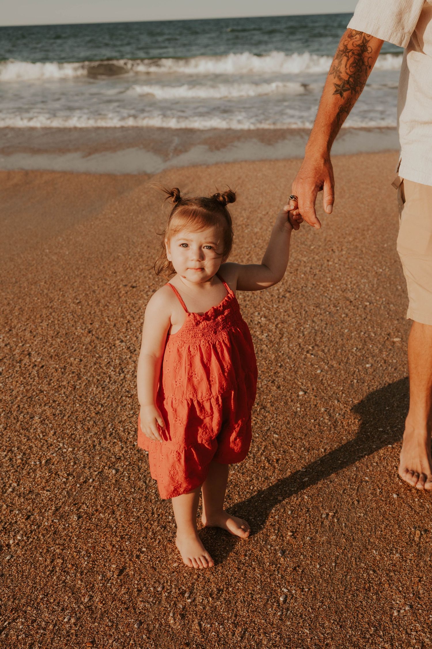 Toddler girl in red dress holds father's hand while standing on sandy beach at golden hour sunset.