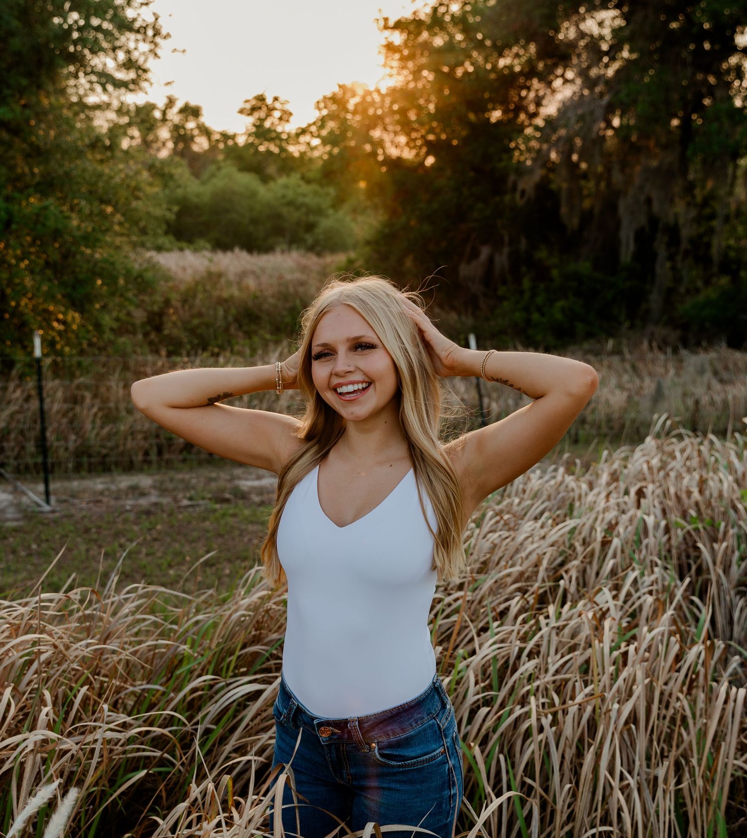 Laughing blonde woman in white bodysuit and jeans standing in tall grass at golden hour.